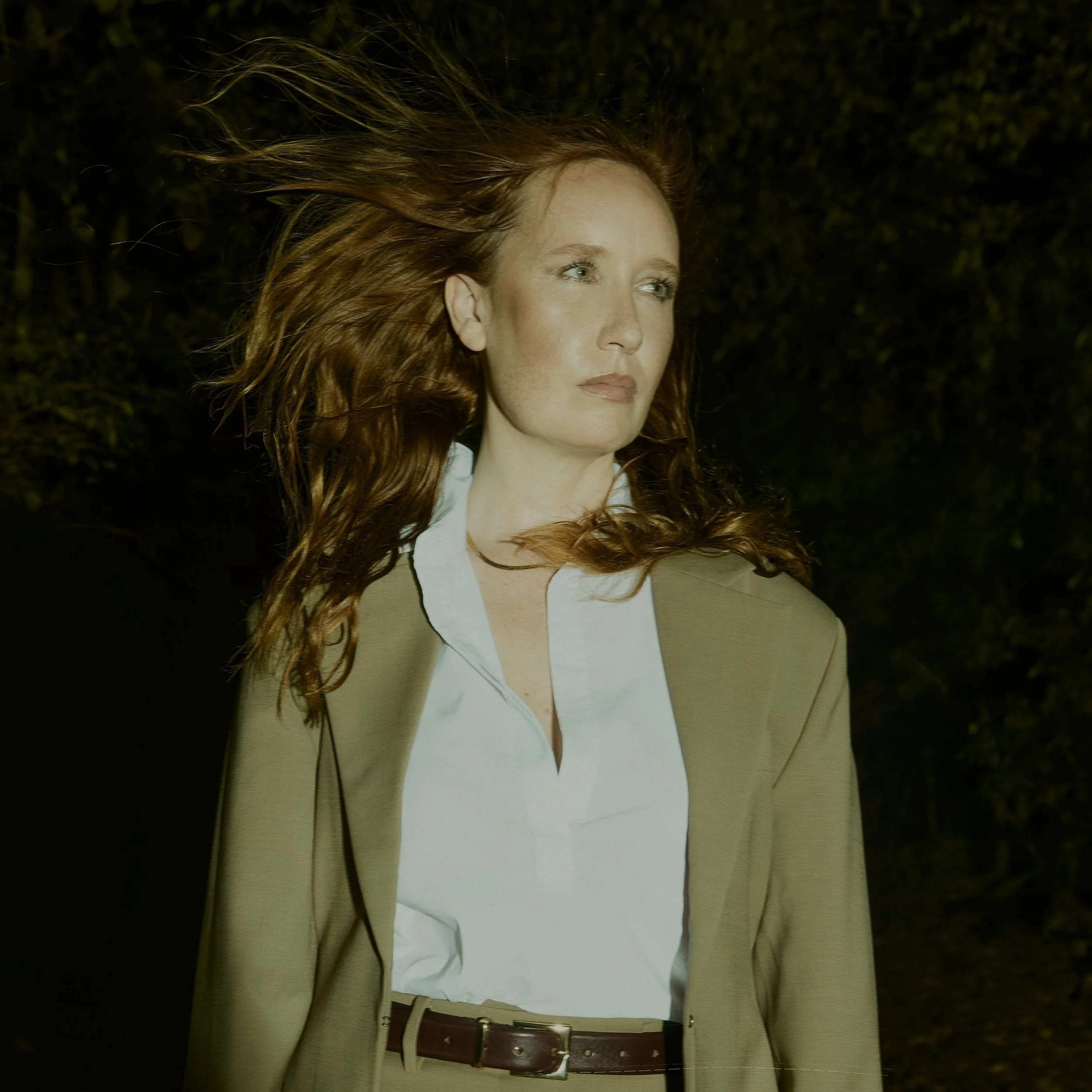 A woman with long red hair standing outdoors at night, wearing a tan suit and white shirt, with wind blowing her hair.