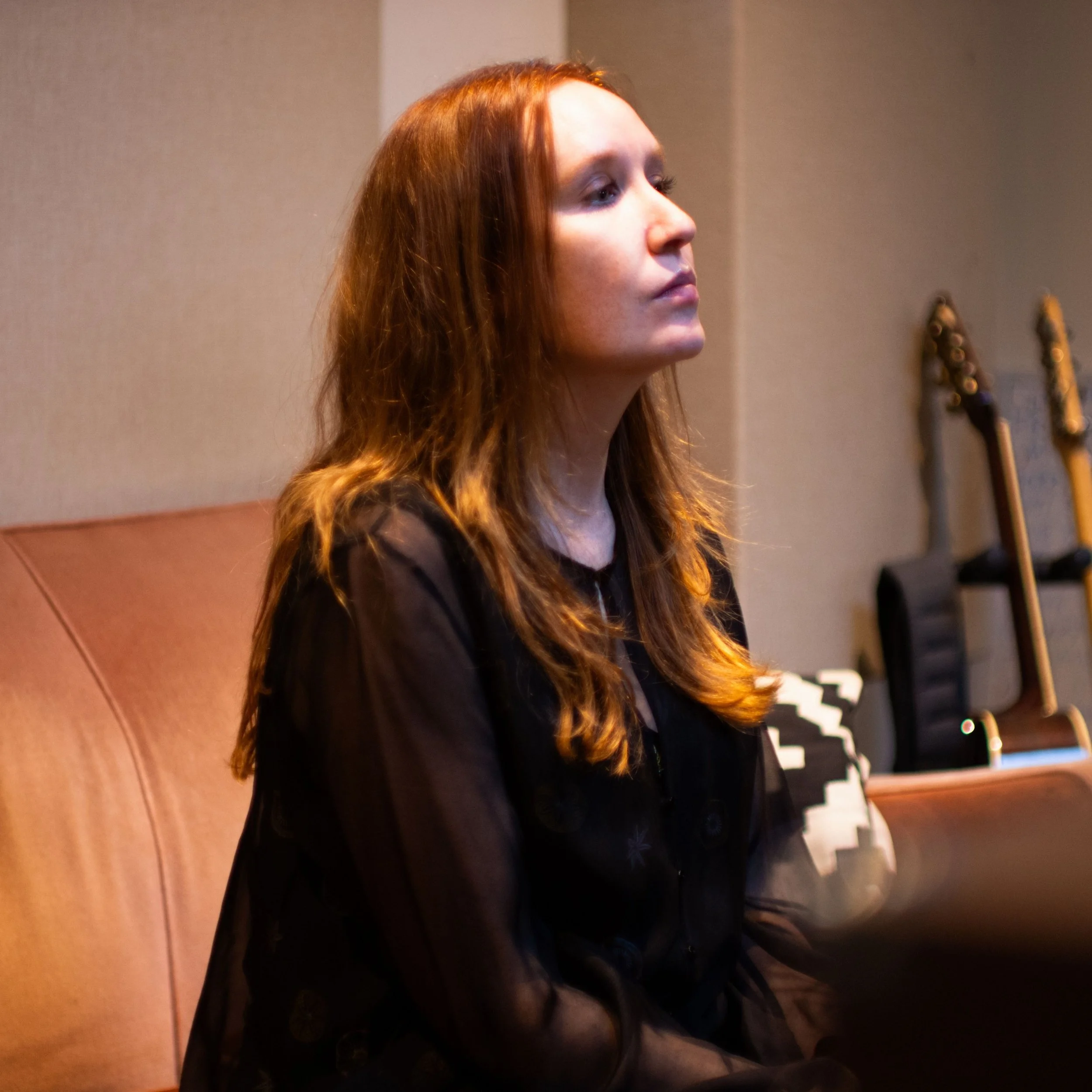 A woman with long, reddish-brown hair, wearing a dark sheer shirt, looking to her left in a music studio.