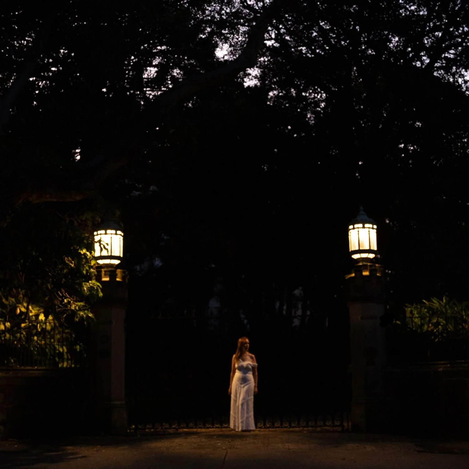 A woman in a white dress standing in front of a black iron gate with two lit lanterns on pillars, surrounded by trees at night.