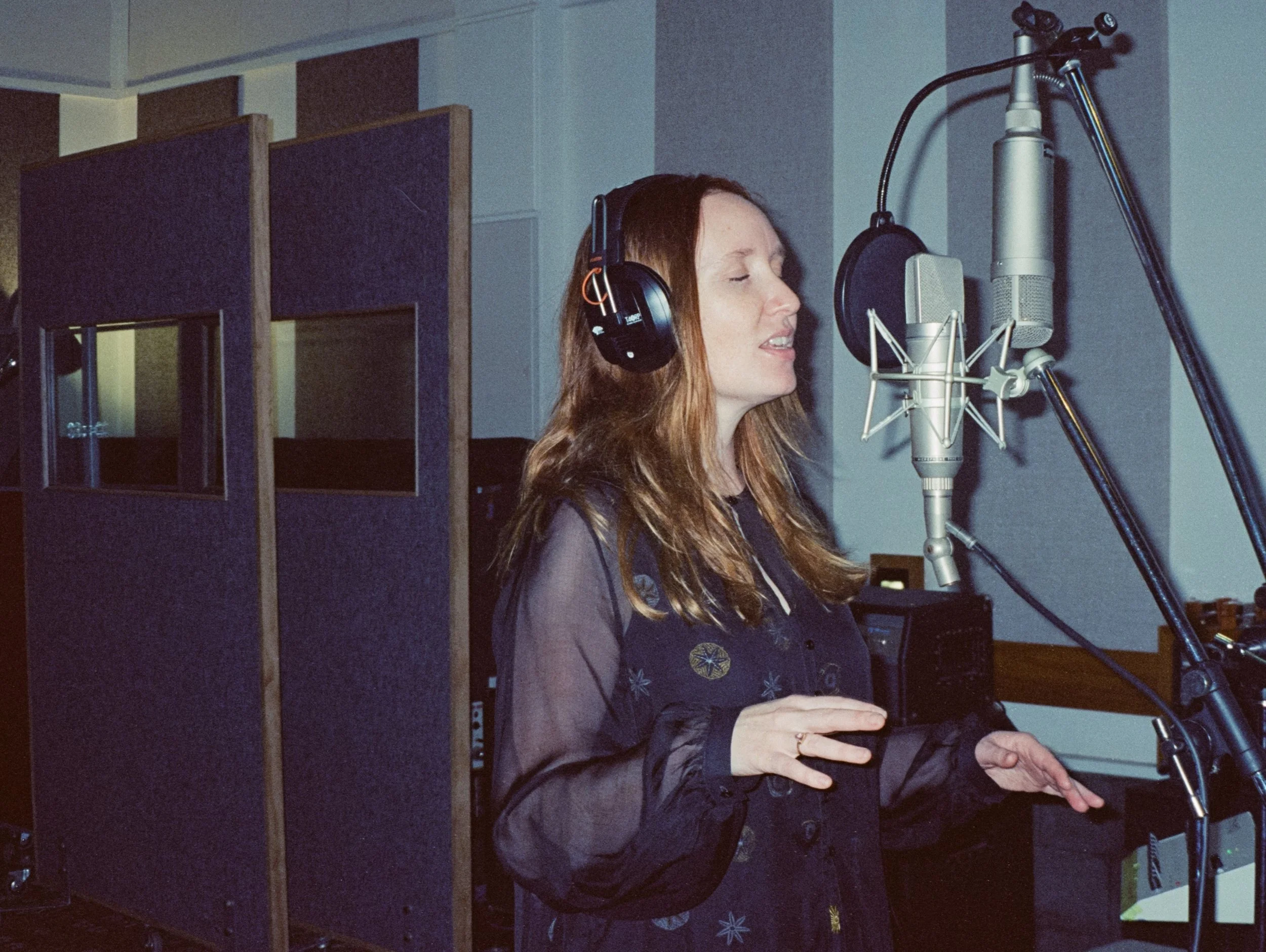 A woman with long, brown hair singing into a microphone in a recording studio, wearing headphones and a dark blouse with floral patterns.