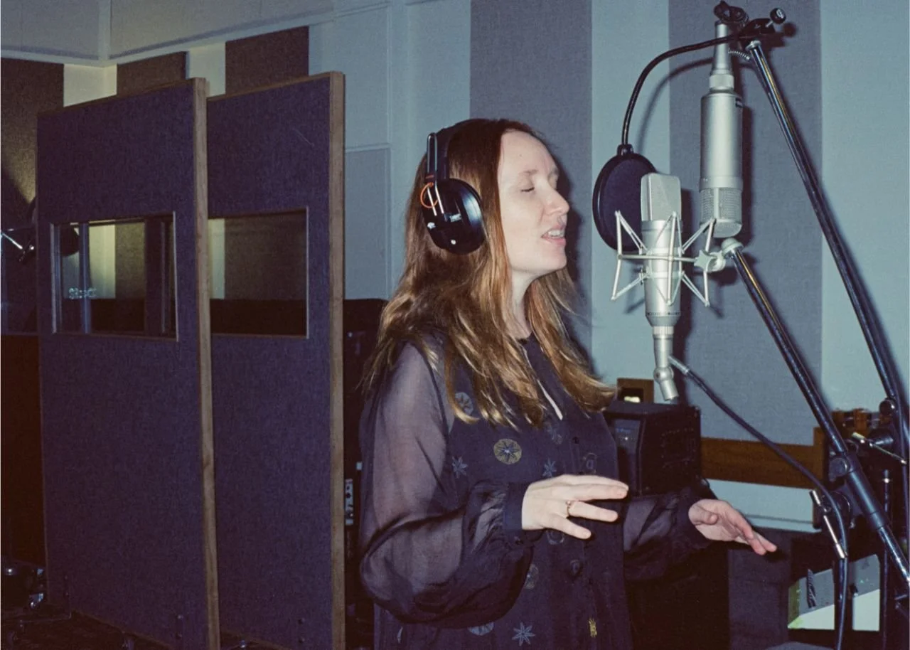 A woman singing into a microphone in a recording studio, wearing headphones.