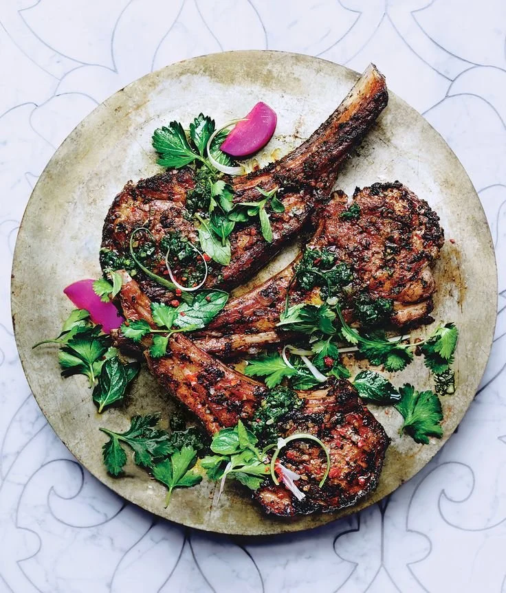 Grilled lamb chops garnished with fresh cilantro, parsley, and microgreens on a round cutting board.