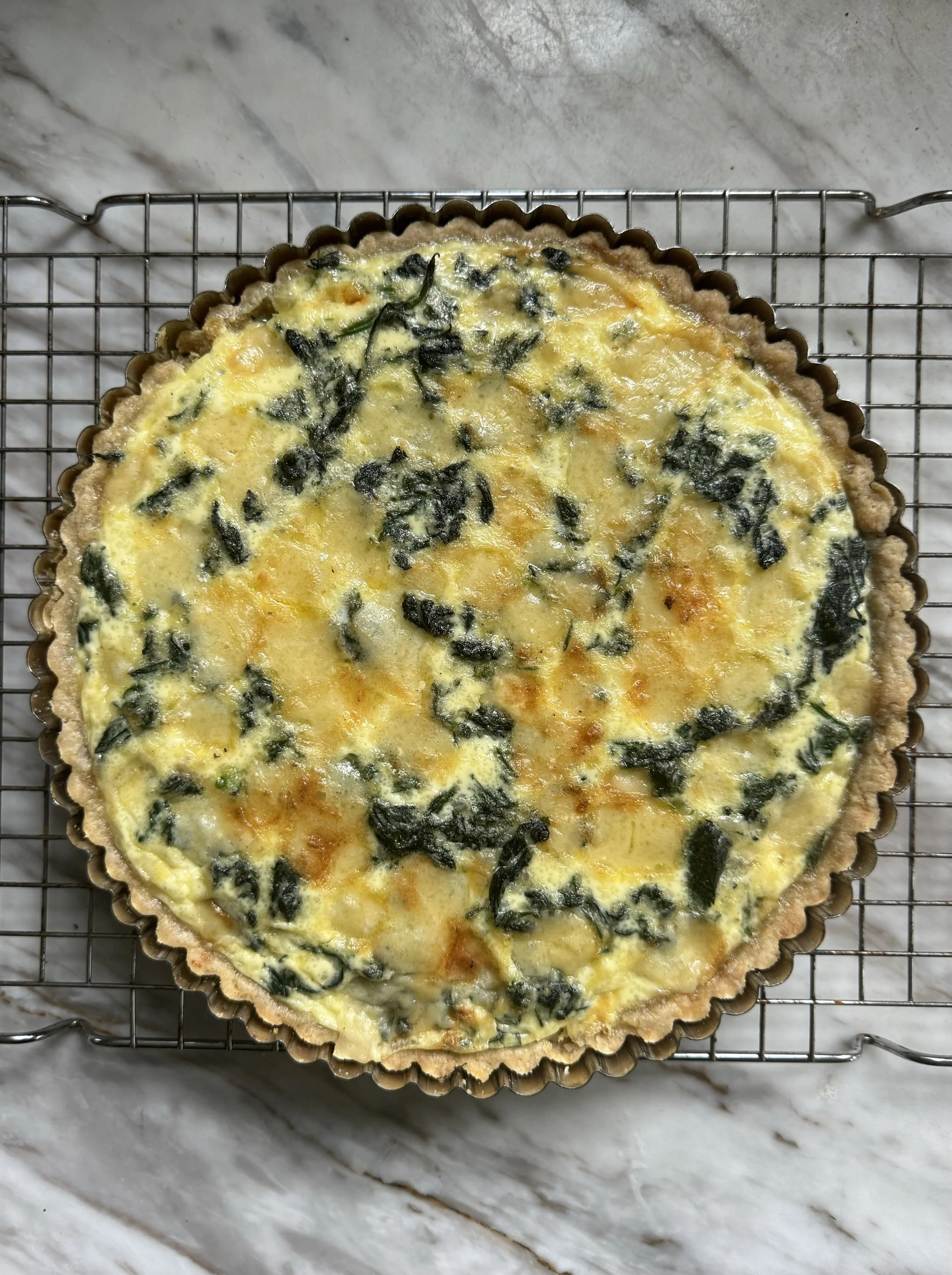 A quiche with spinach and cheese on a wire cooling rack on a marble surface.