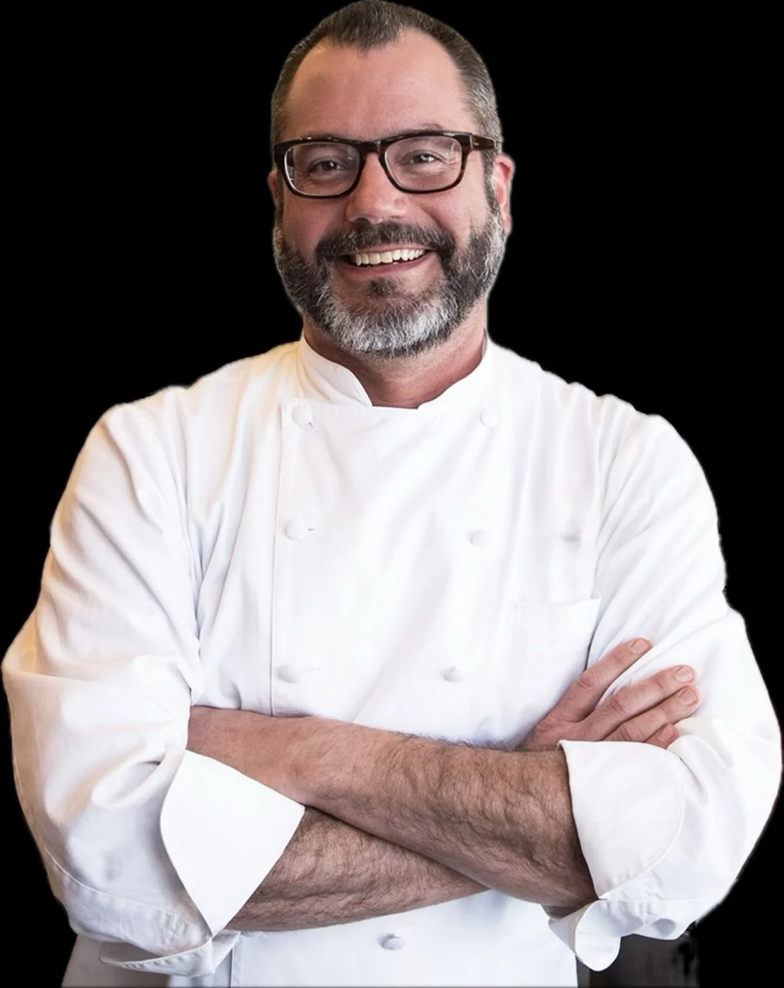 Smiling male chef with glasses, grey beard, wearing a white chef's coat, standing with arms crossed against a black background.