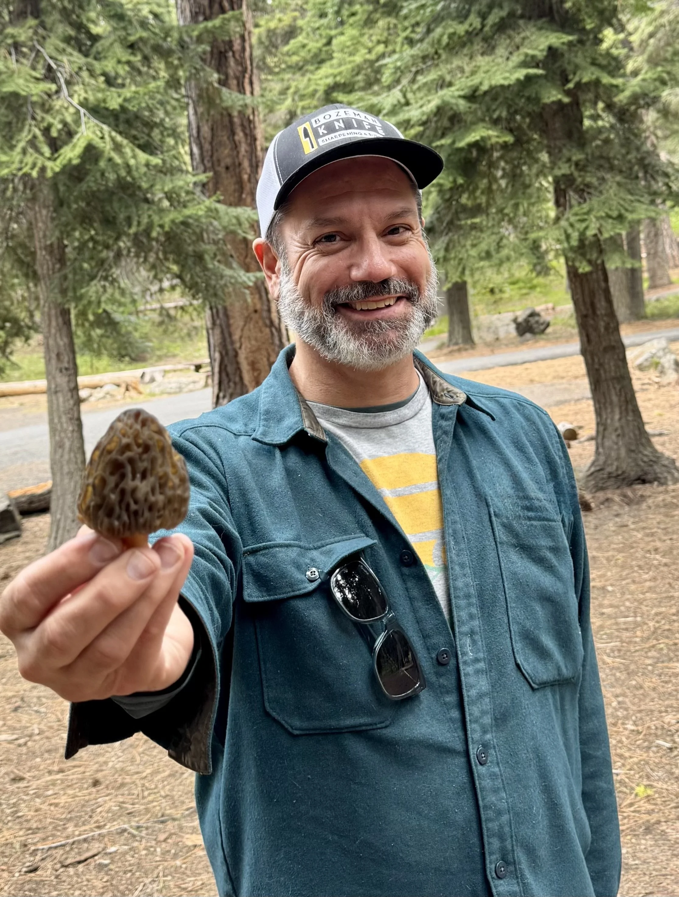 A man outdoors in a forest holding a small brown mushroom towards the camera. He is wearing a dark green shirt, a cap, and sunglasses hanging from his shirt pocket, smiling at the camera.