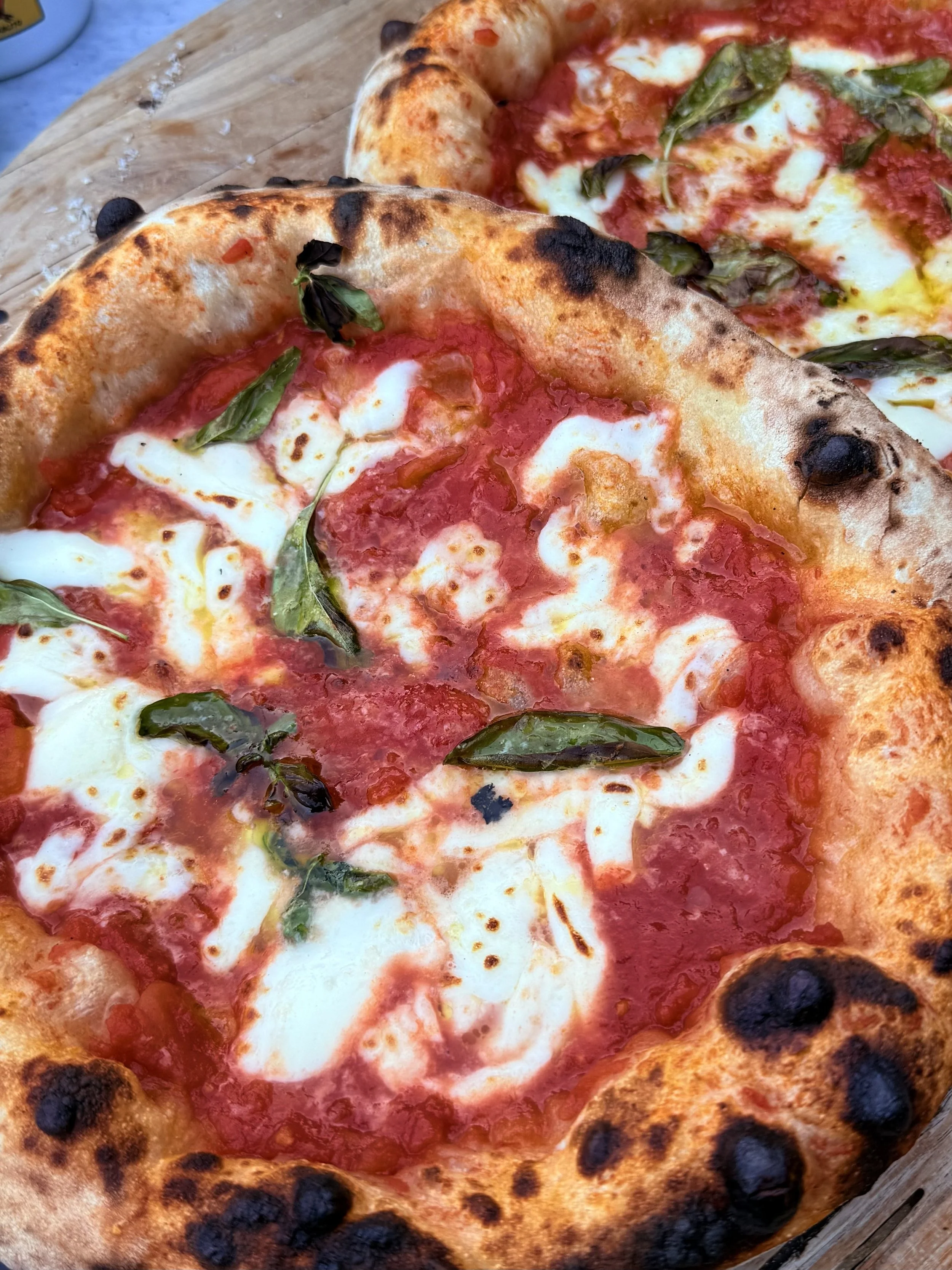 Two Margherita pizzas with melted cheese, tomato sauce, basil leaves, and a slightly charred crust on a wooden surface.