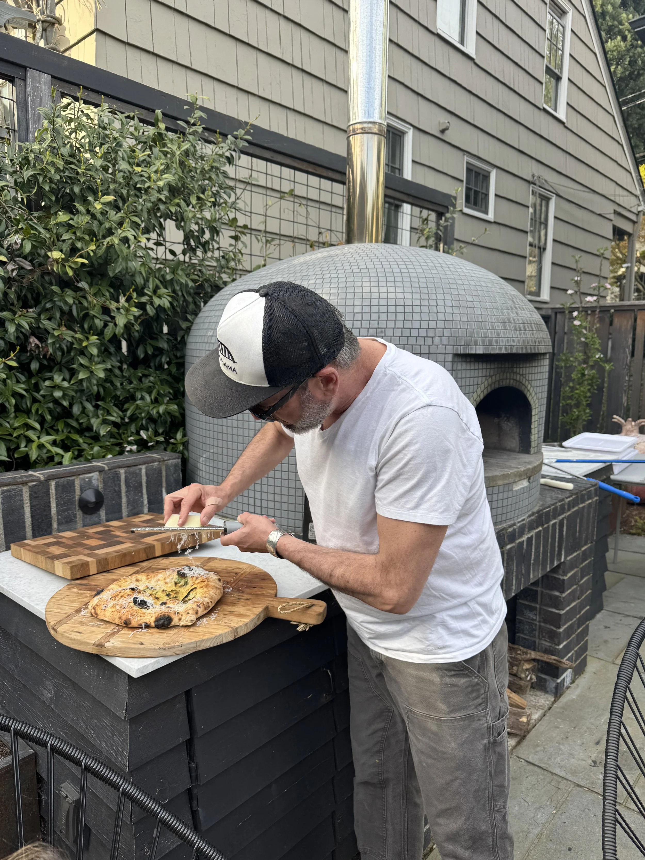 A man wearing a baseball cap, sunglasses, and a white t-shirt is slicing a pizza on a wooden cutting board outdoors near a brick oven, with greenery and a house in the background.
