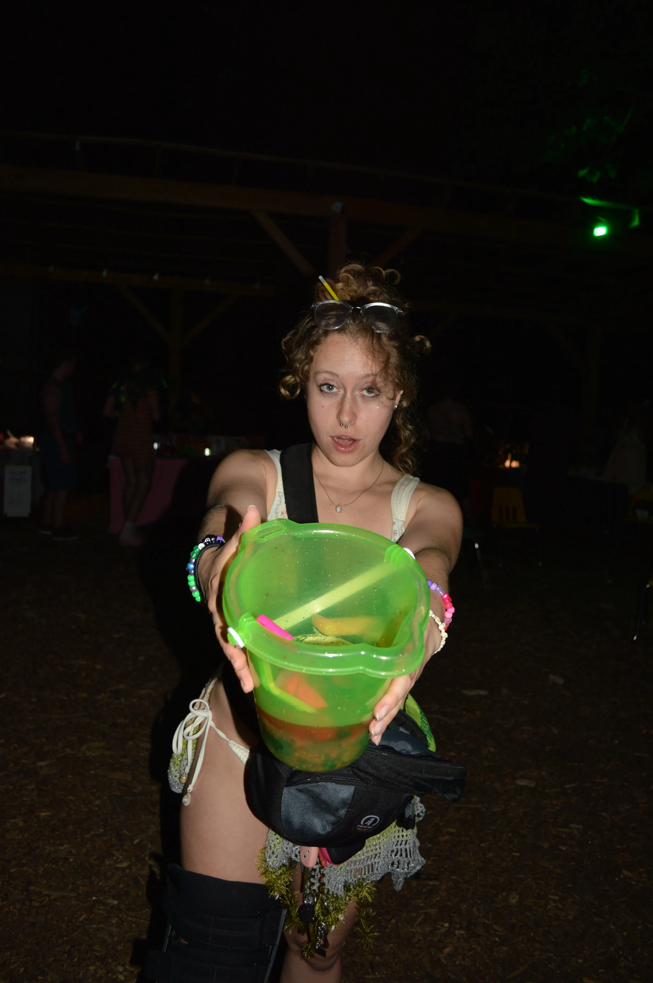 Young woman with curly hair, sunglasses on her head, and a septum piercing, holding a green bucket with a yellow toy inside, at night in an outdoor setting with others in the background.