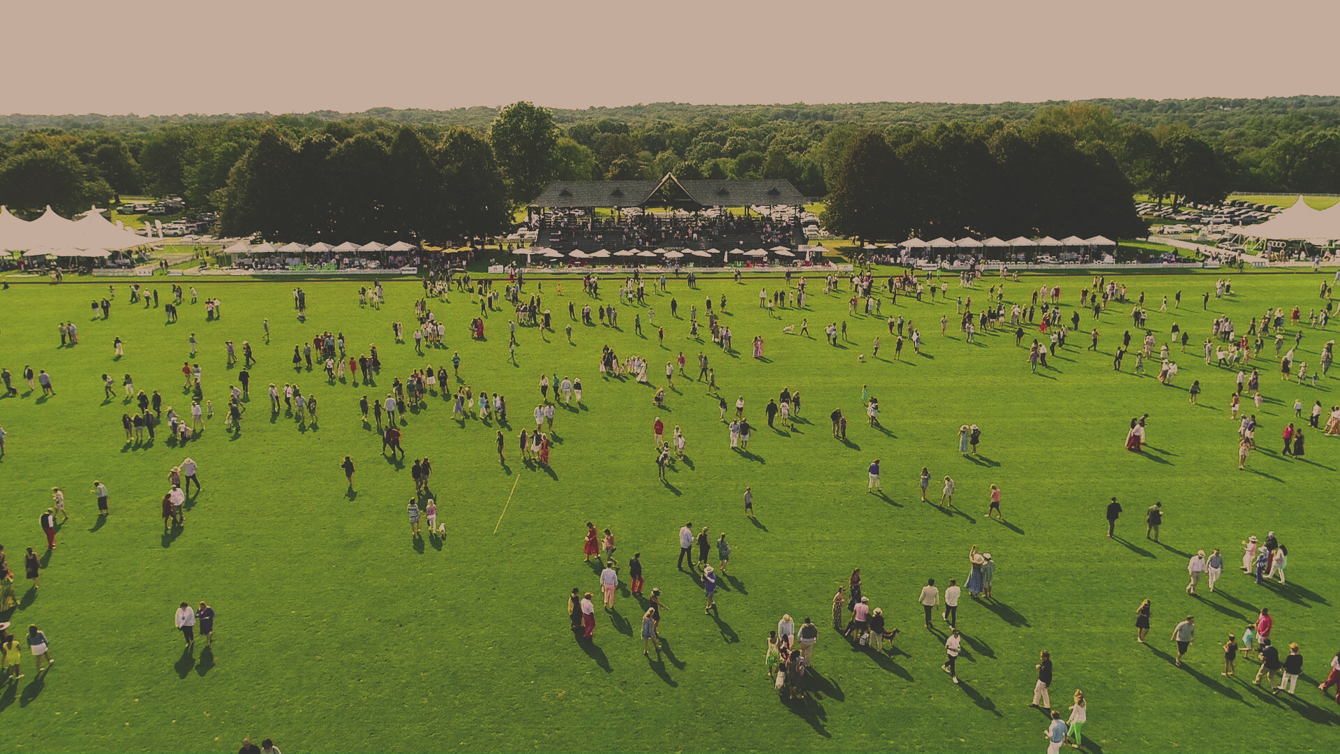 A large outdoor event with many people walking on a green lawn, tents and a stage in the background, and a wooded area beyond.