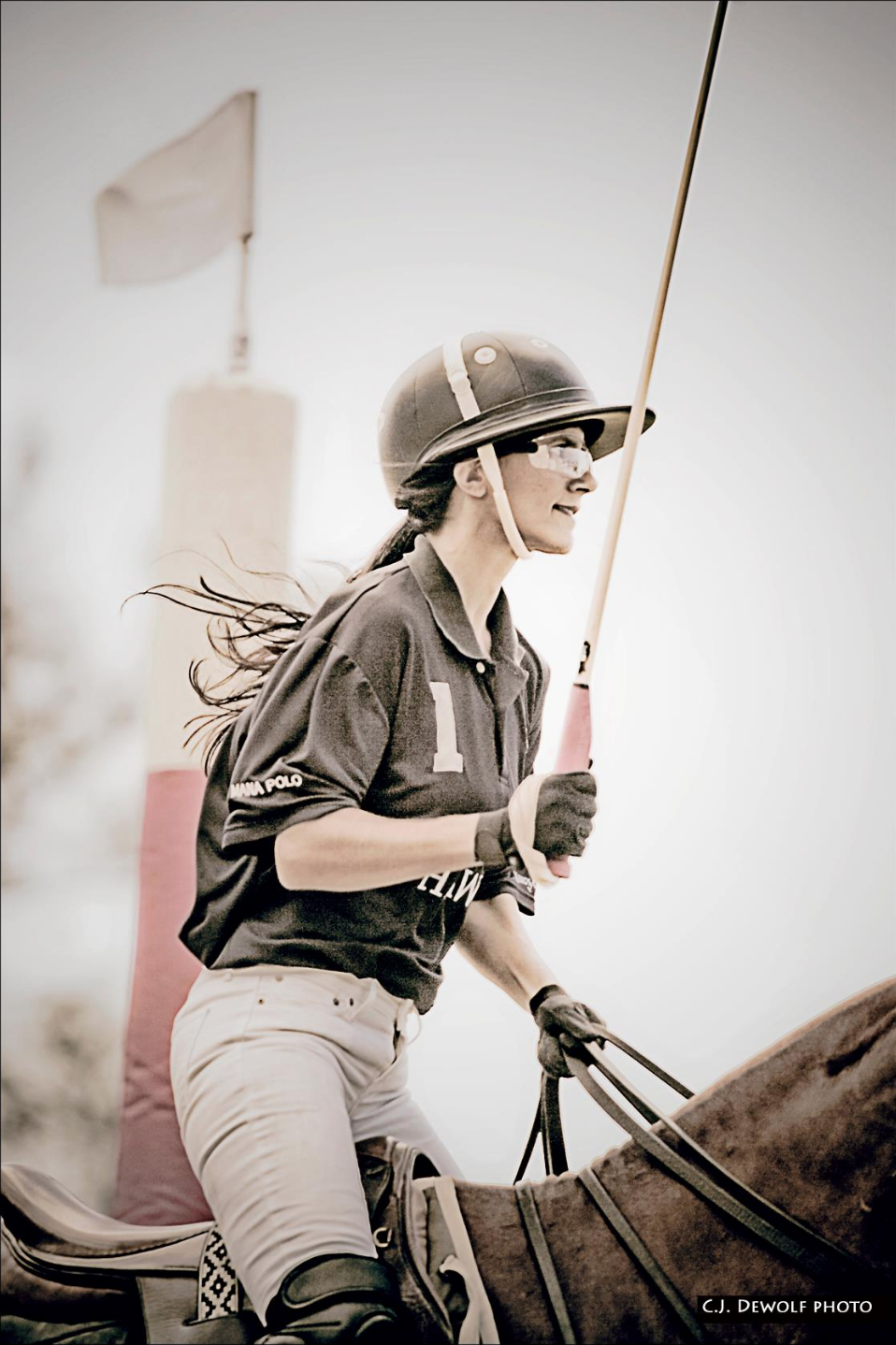 A woman riding a horse, dressed in equestrian attire, holding a polo mallet, with her hair flowing in the wind, wearing a helmet, riding gloves, and sporting a smile.