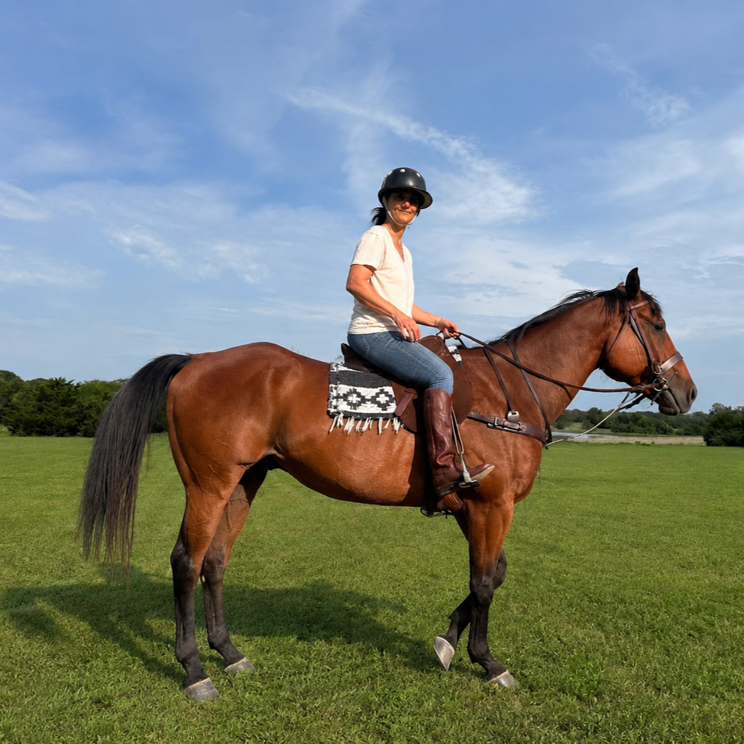 Woman riding a brown horse in a grassy field under a partly cloudy sky, wearing a black helmet, sunglasses, a white shirt, and jeans.