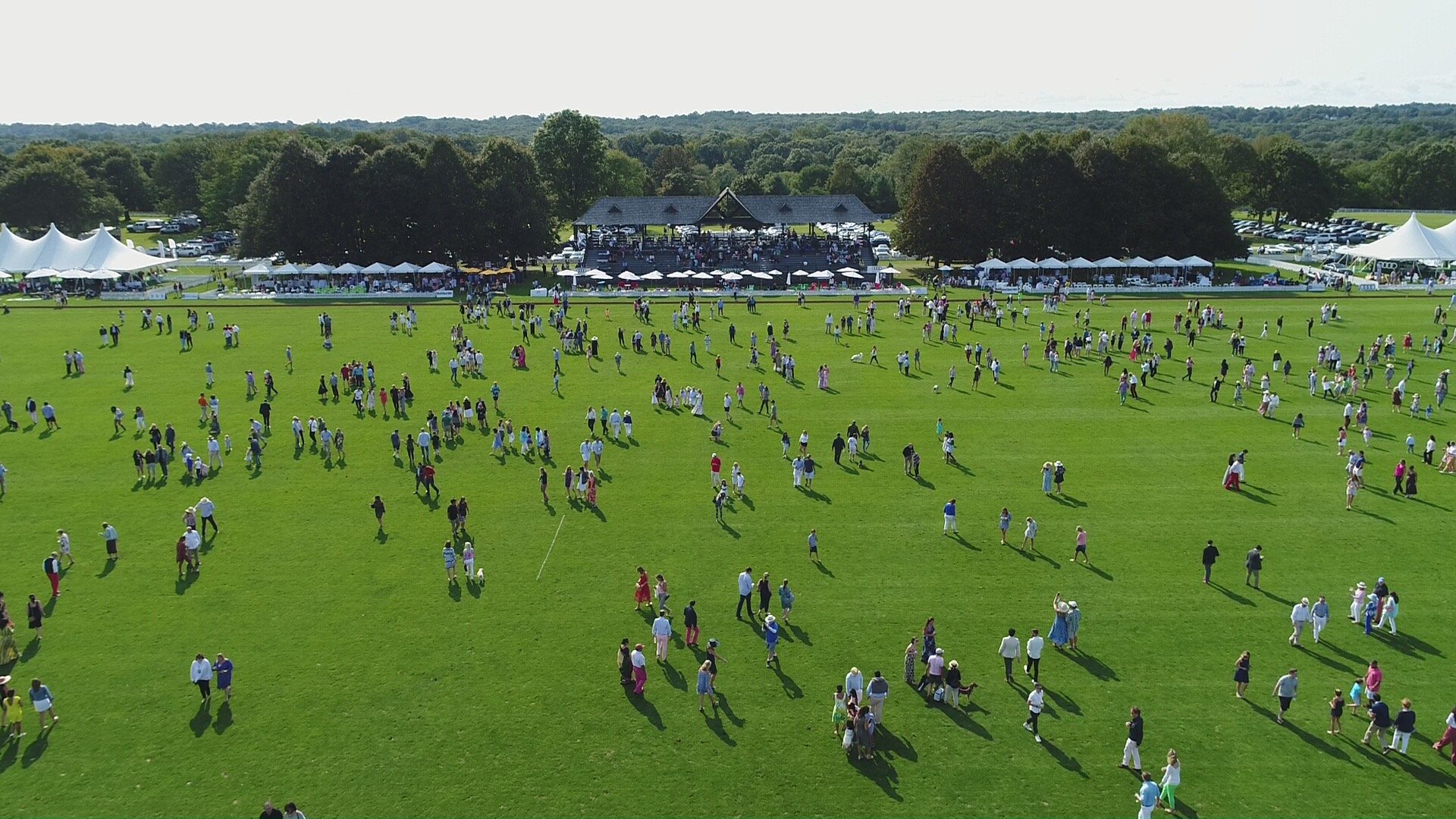 A large outdoor event with many people walking on a green field, tents, and a stage with seating under trees. Bright sunny day with clear skies.