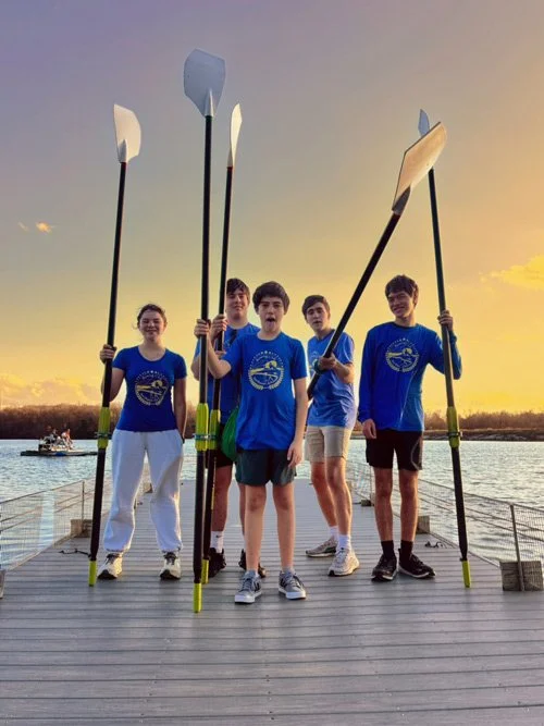 Four teenagers standing on a dock holding paddles, wearing matching blue T-shirts with a logo, during a sunset at a lake.