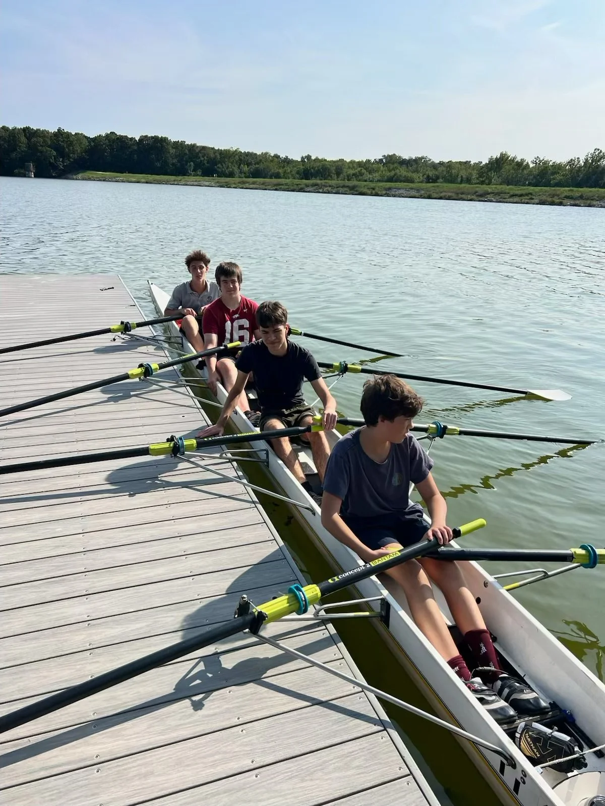 Four boys sitting in a rowing boat docked at a wooden pier on a calm body of water, with a green treeline in the background, preparing for rowing practice.