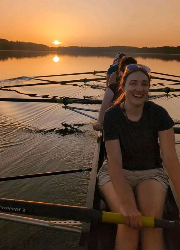 A group of people in a boat on a calm lake at sunset, with the sky orange and the water reflecting the sunset. The person in the front is smiling and holding an oar, wearing a black t-shirt and sunglasses on her head.