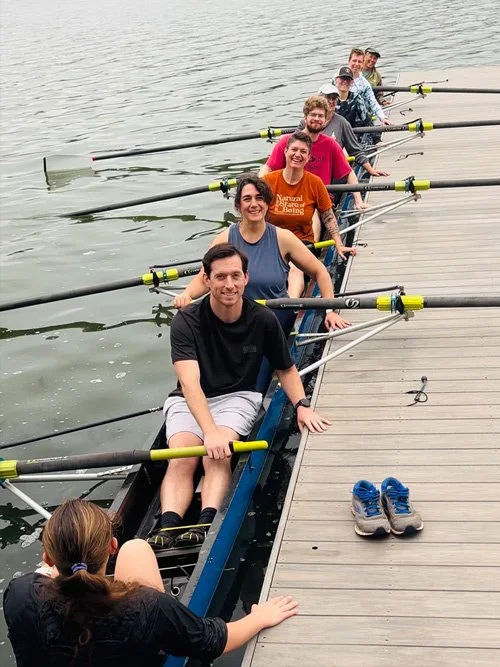 Group of people in a rowing boat along a dock, with shoes next to the dock, on a body of water.