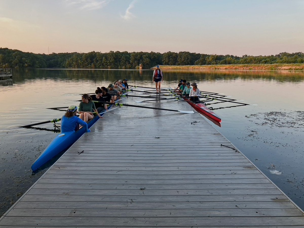 Group of people with paddles and racing shells on a dock, preparing to row on a calm lake during sunset.