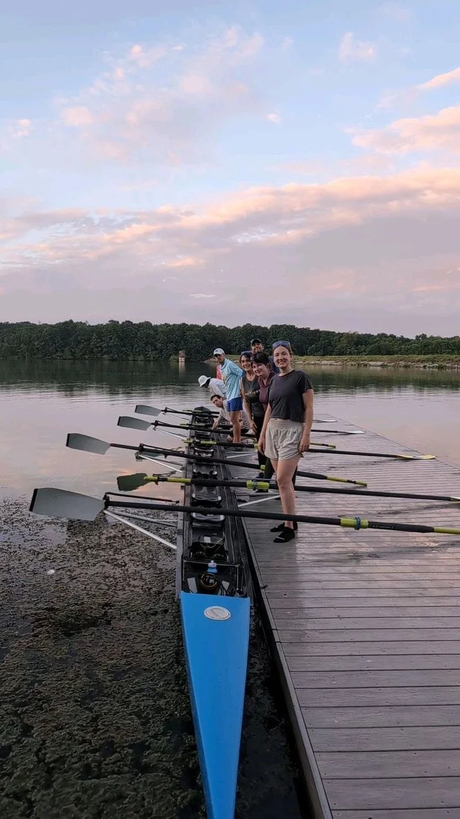 Group of five people standing on a dock with rowing shells and oars, preparing to row on a calm lake during sunset.