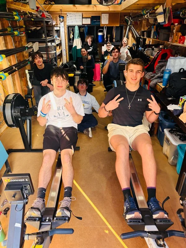Group of young people in a cluttered gym or storage room, some sitting on rowing machines and others standing behind, surrounded by shelves filled with sports equipment, bags, and miscellaneous items.