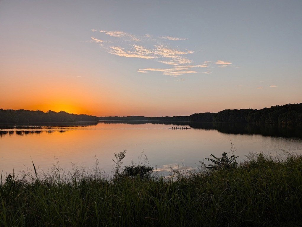 Sunset over a calm river with a small boat and people rowing in the distance, surrounded by trees and grassy shoreline.