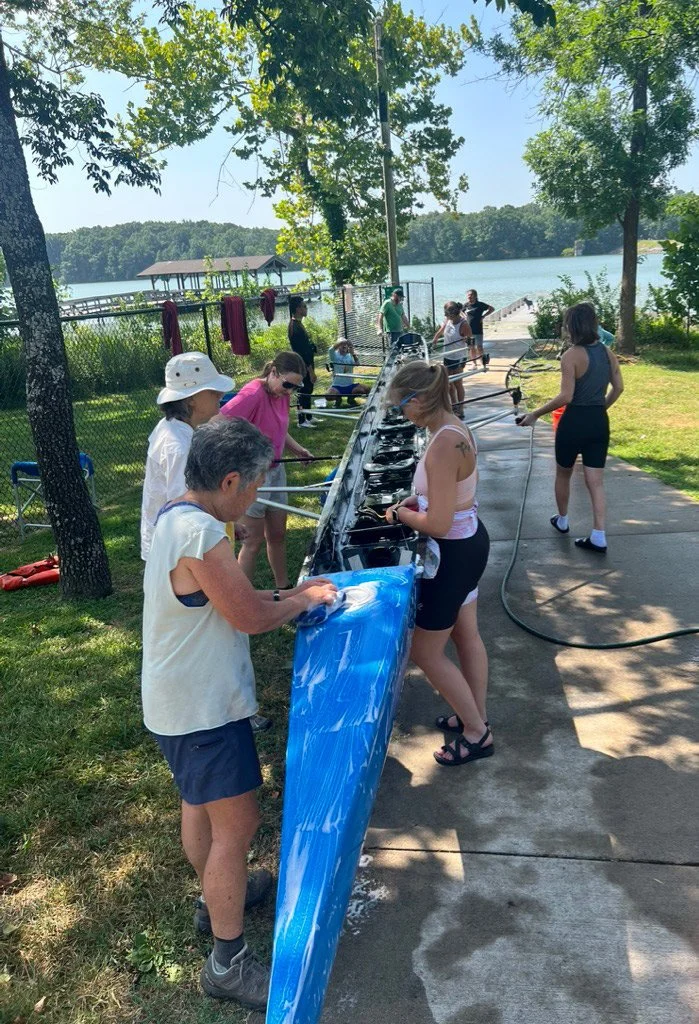 People cleaning and washing kayaks near a lake on a sunny day, with trees and a dock in the background.