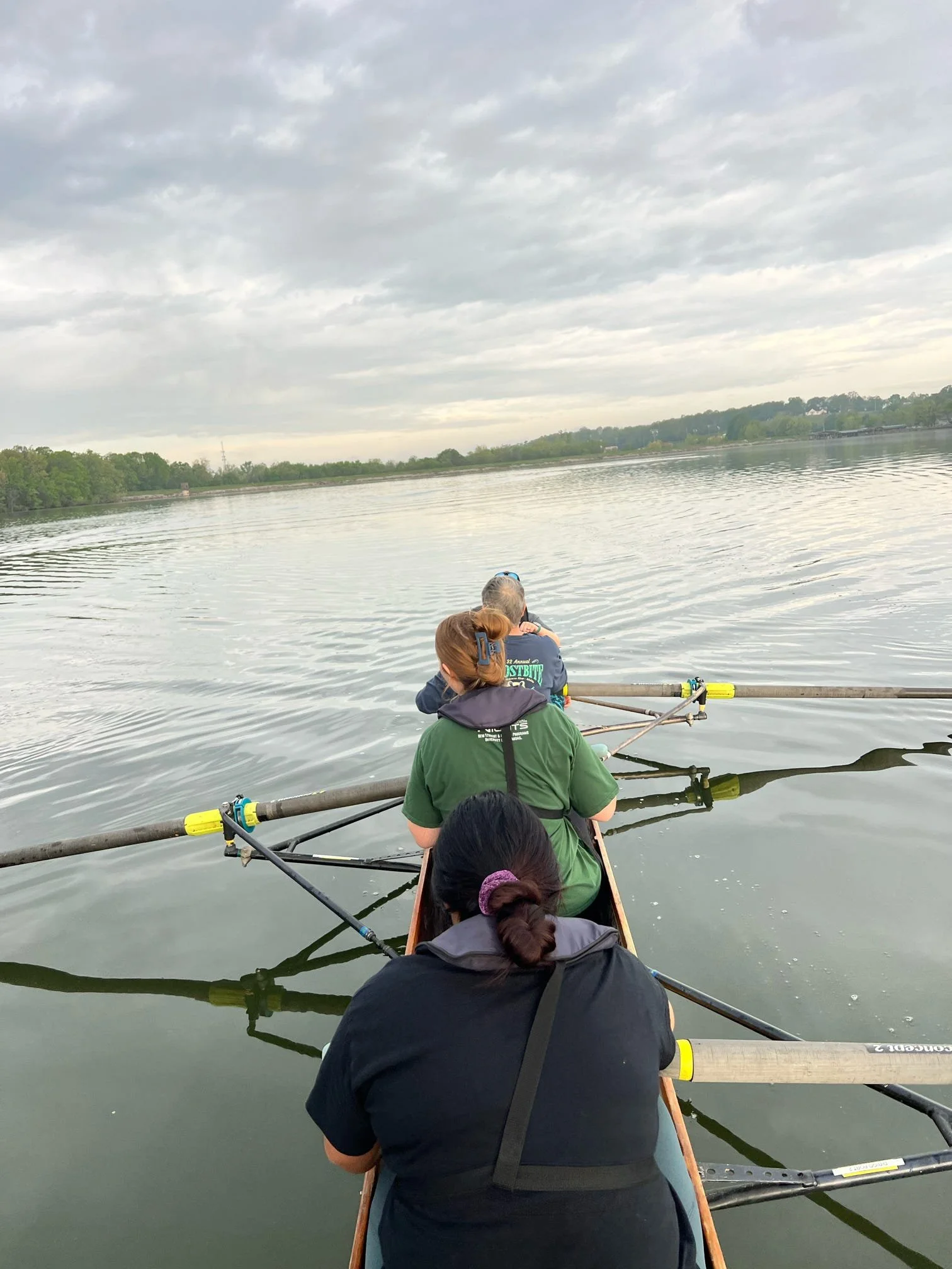 People in a boat rowing on a calm river under a cloudy sky, with trees and a distant shoreline in the background.