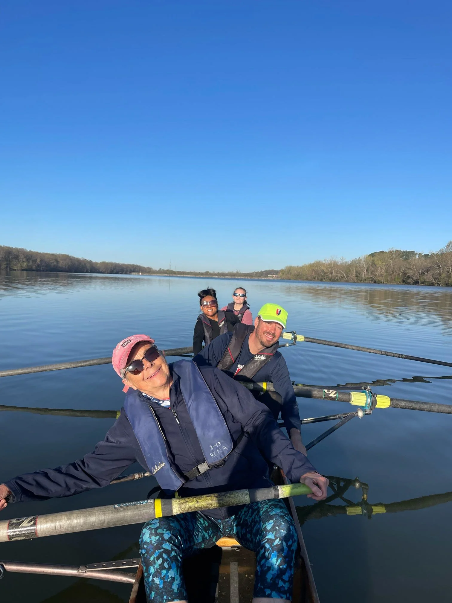 A group of five people smiling while rowing a boat on a calm lake under a clear blue sky, with trees lining the far shore.