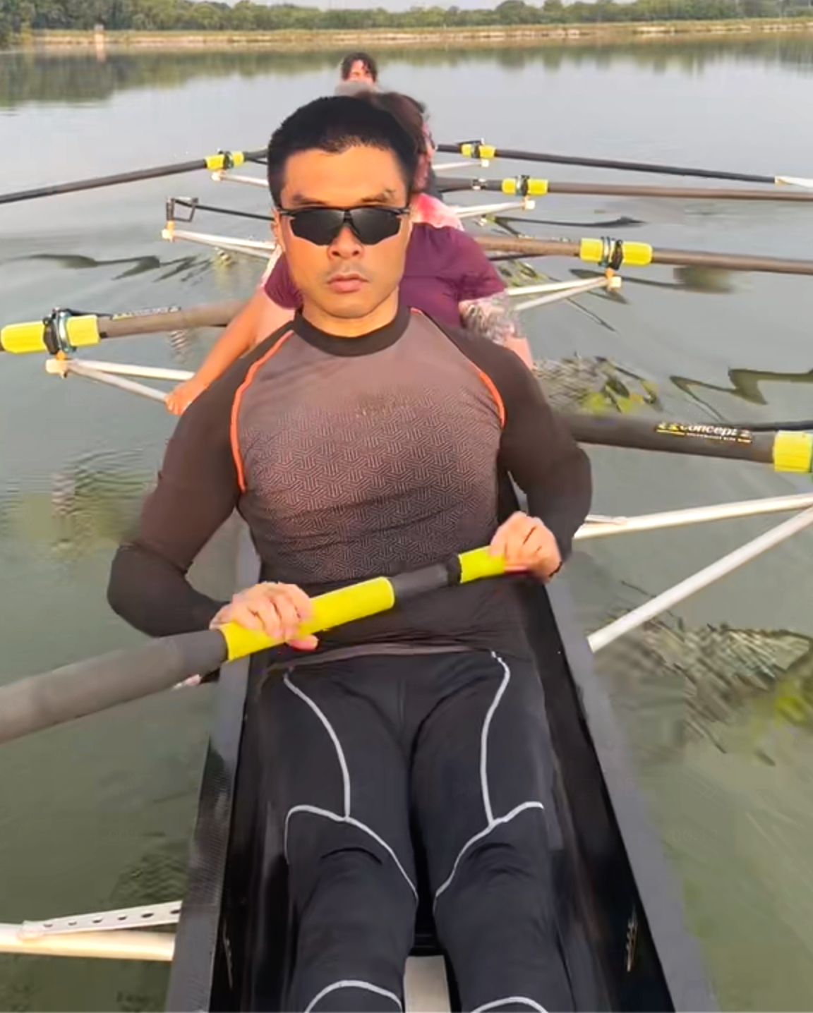 People rowing a boat on a calm lake, with the man in the front wearing sunglasses and a black and gray athletic shirt.