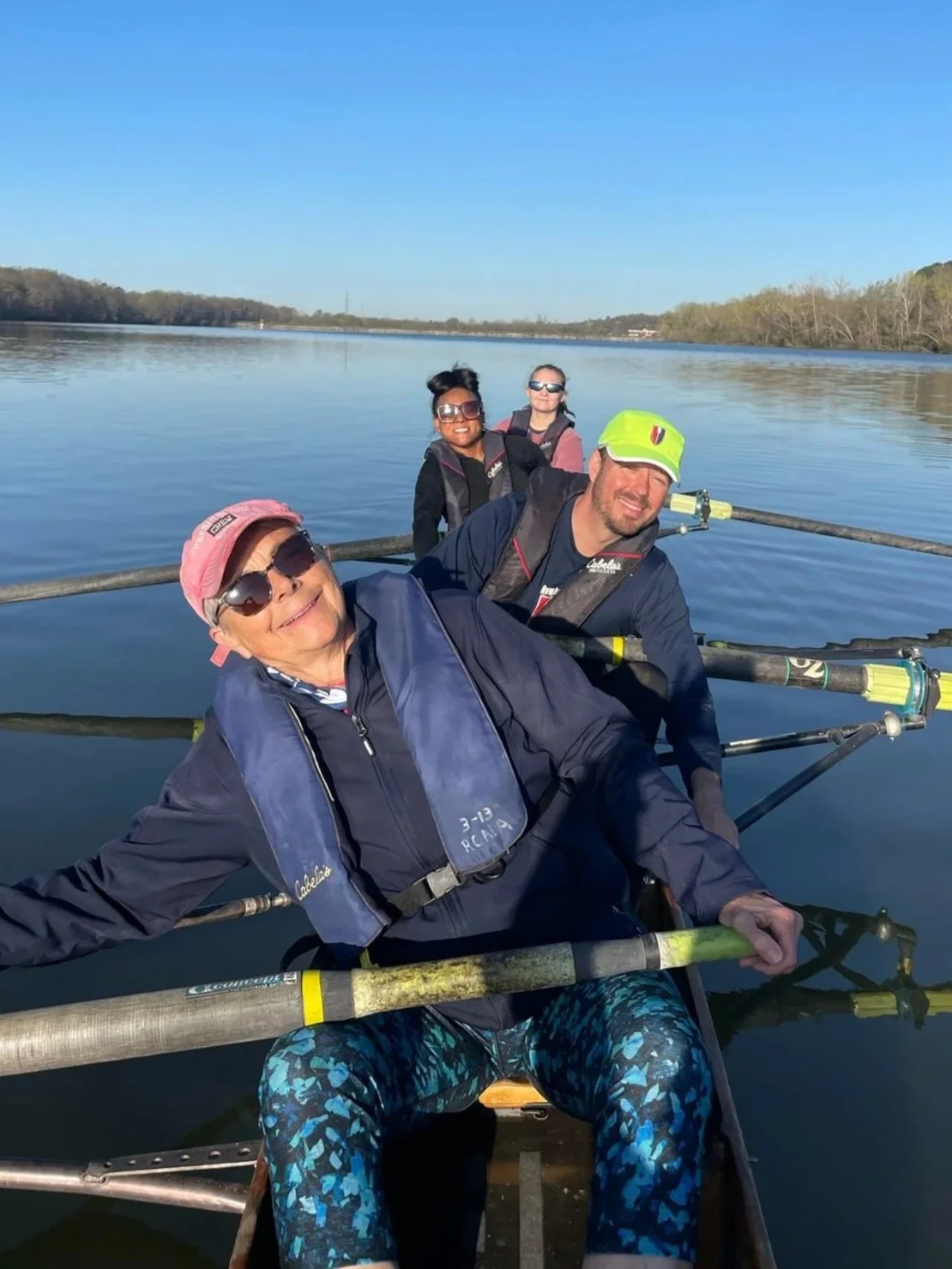 Four people rowing a boat on a calm river on a sunny day, with trees in the background.