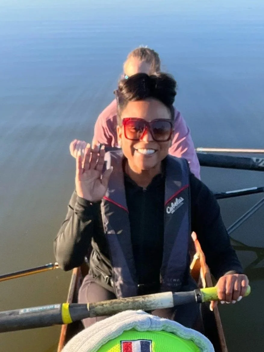 Two women kayaking on calm water during sunset, one smiling and waving, wearing sunglasses and a life jacket.