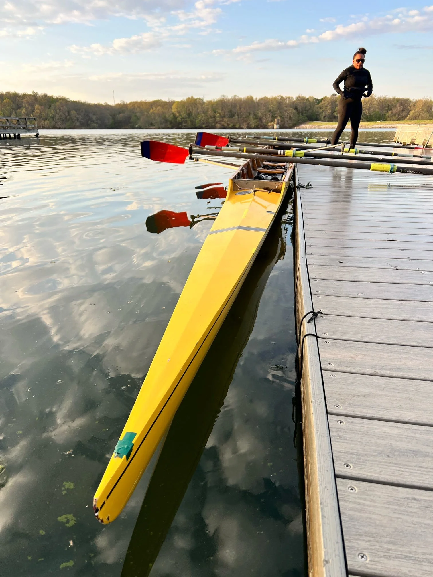 A woman standing on a dock next to a yellow racing shell with oars, with water and trees in the background under a partly cloudy sky.