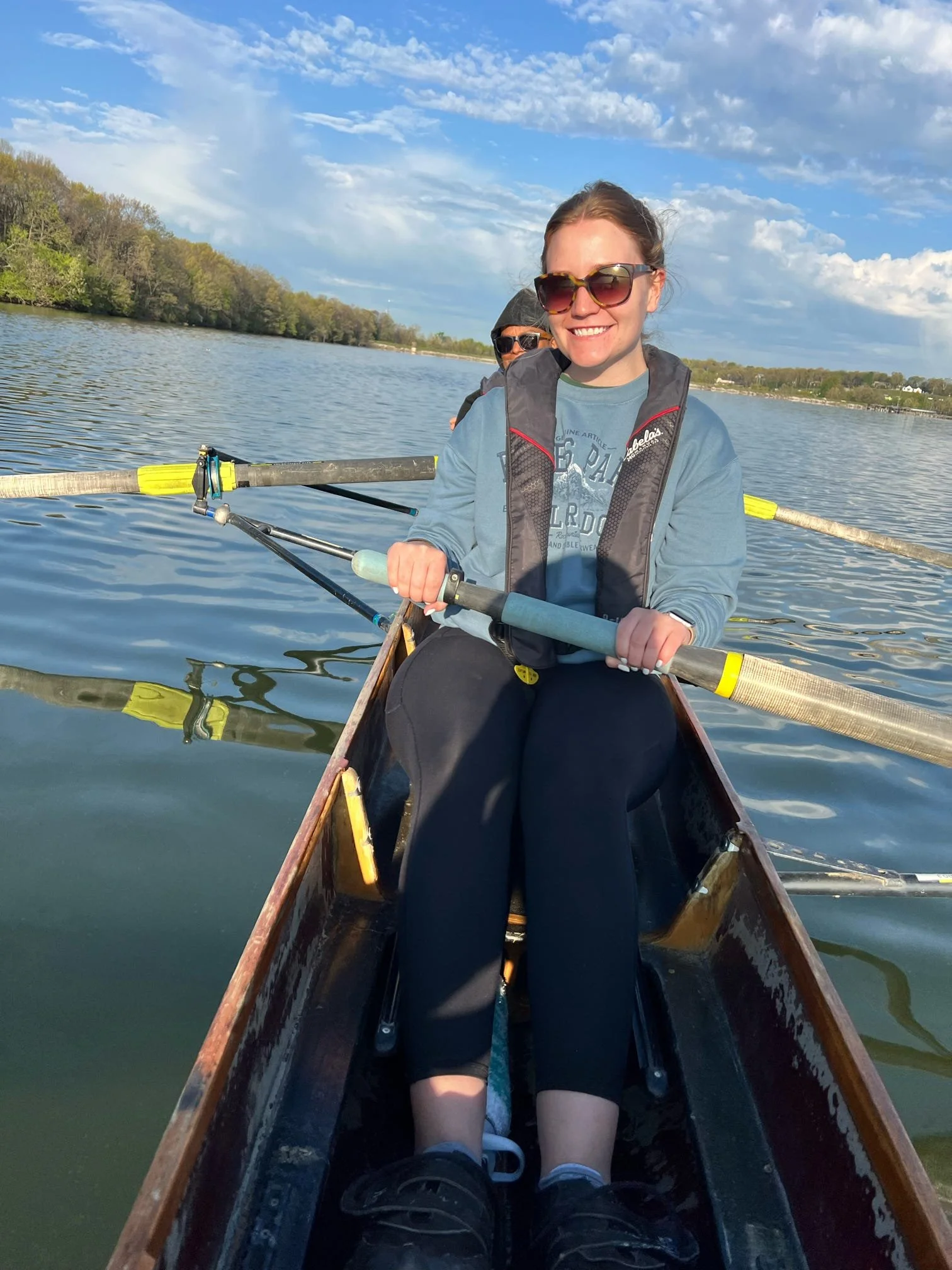 A woman smiling while rowing a boat on a calm lake with a person in the background, trees along the shore, and a partly cloudy sky.