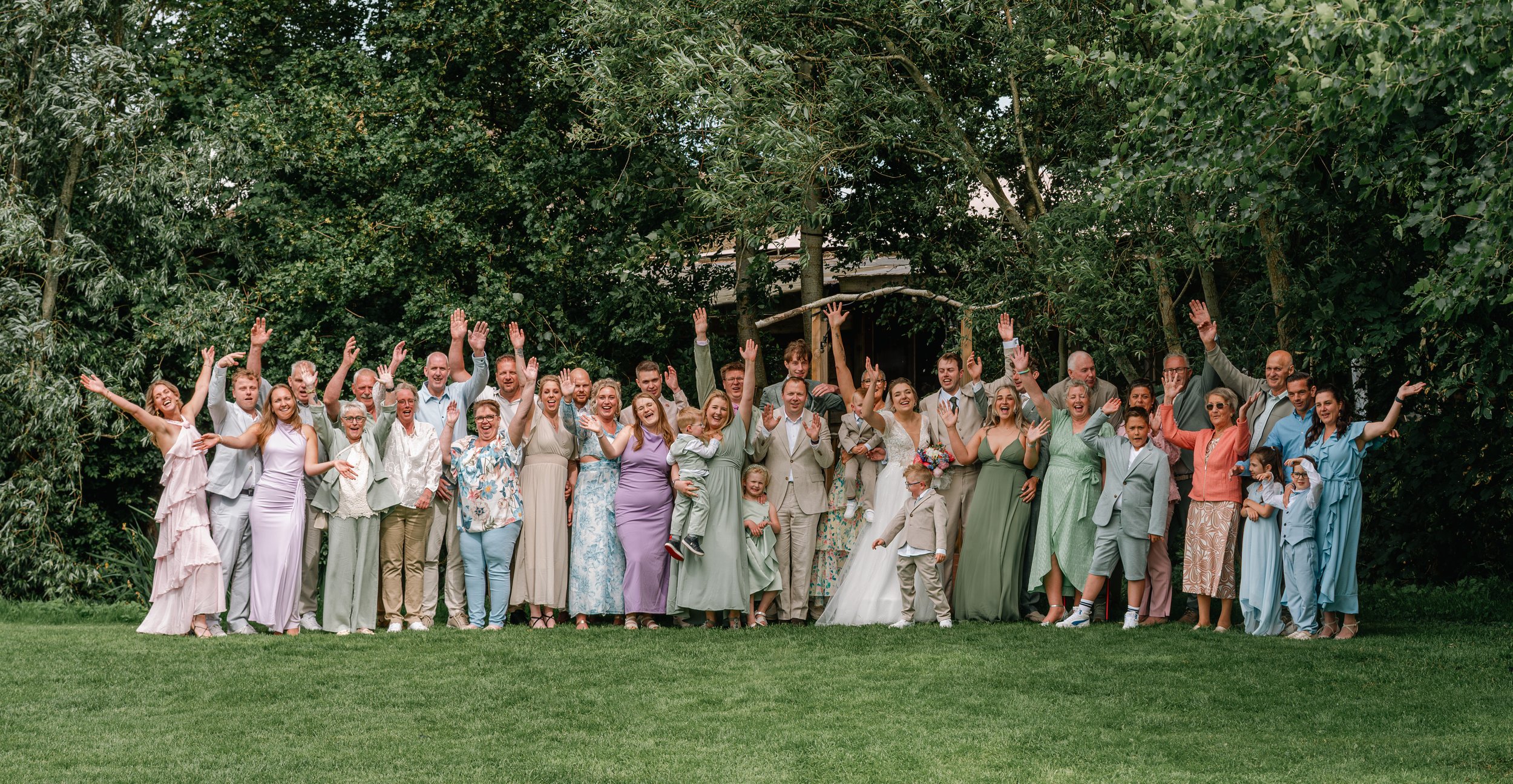A large group of people, including adults and children, gathered outdoors on a lawn, celebrating a wedding. They are dressed in formal attire and are waving and smiling, with a lush green background of trees.