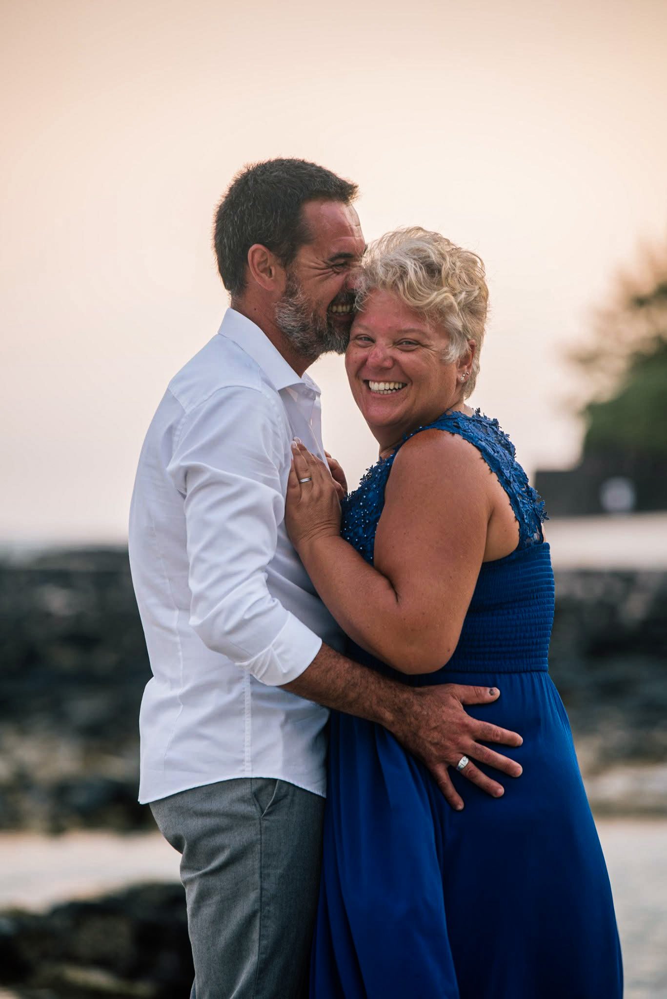 A happy couple, a man and a woman, embracing and smiling outdoors at sunset. The man has gray hair and a beard, wearing a white shirt, and the woman has short blonde hair, wearing a blue dress. They are close to each other, showing affection and joy.