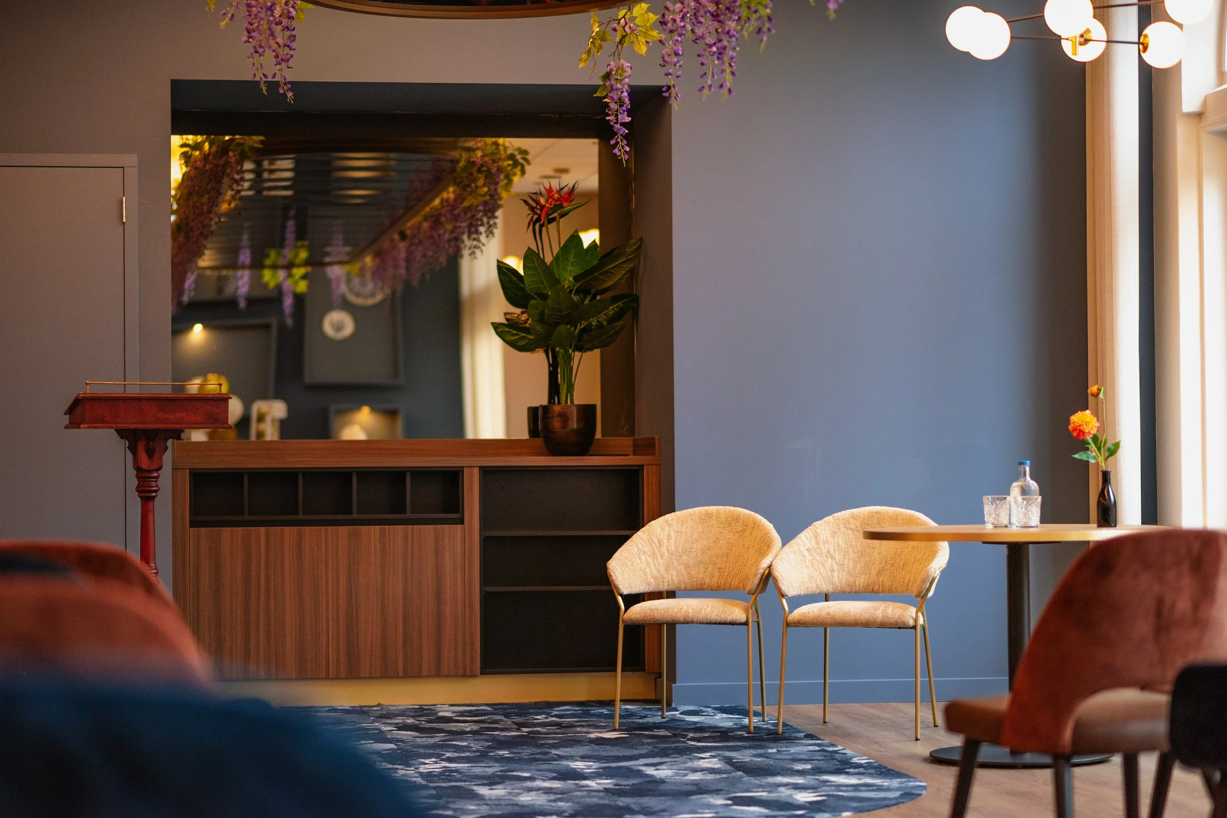 Interior of a room with a blue wall, decorated with purple hanging flowers, hanging white globe lights, a wooden sideboard with a potted plant, a small table with a glass bottle and a flower, and beige chairs.