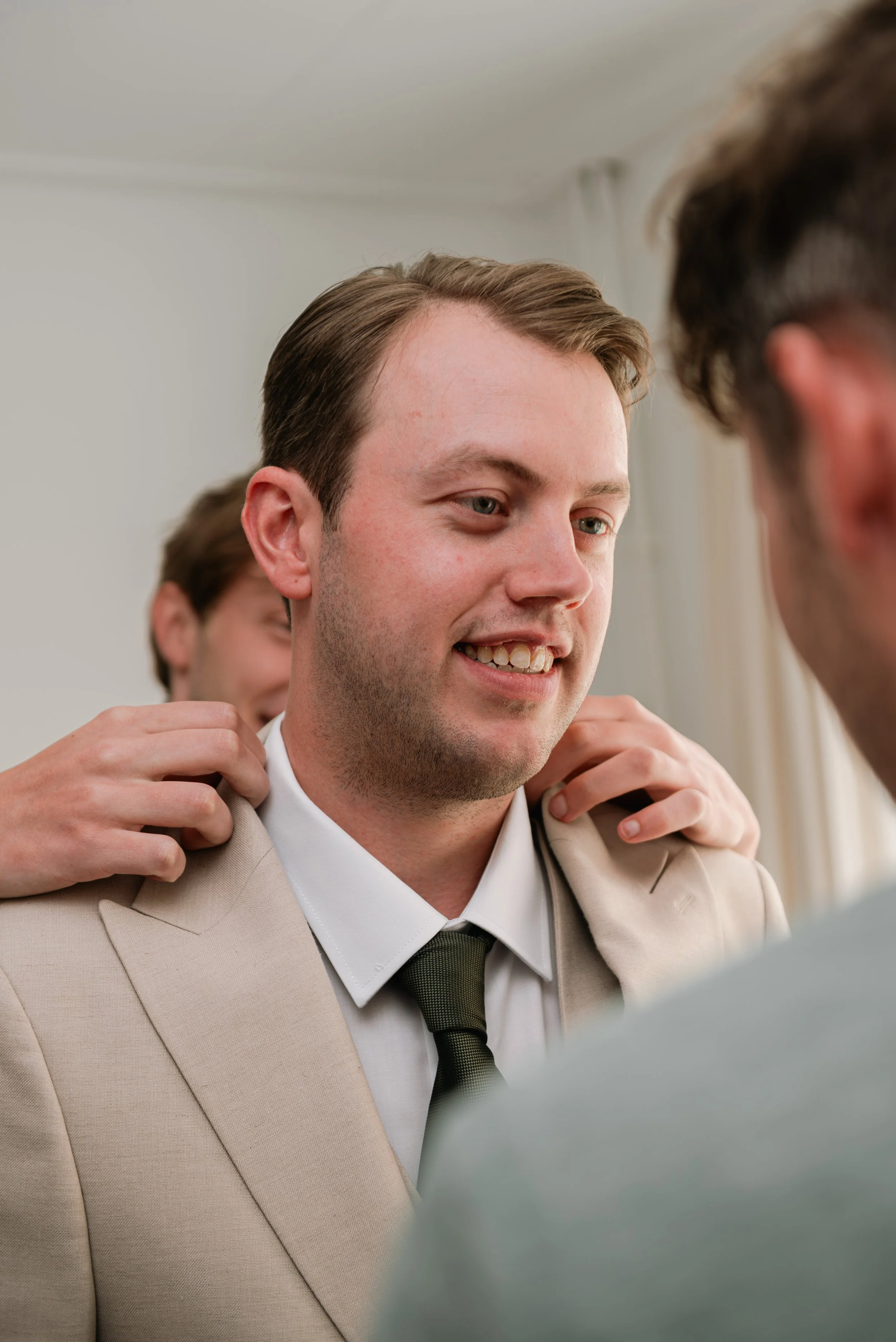 Young man in beige suit smiling as others help him with his tie, in a formal setting.