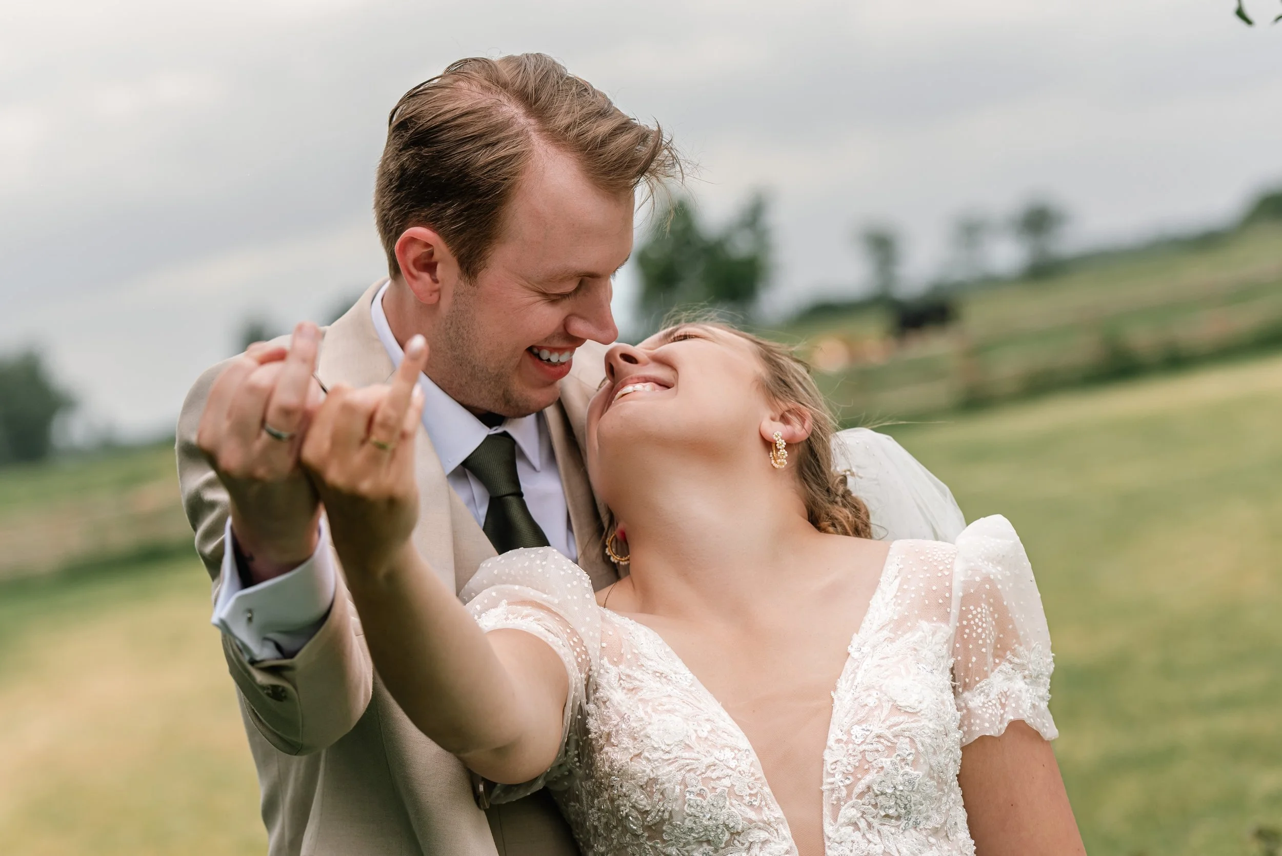 A joyful couple, with the man in a tan suit and the woman in a white lace dress, sharing a dance outdoors on a cloudy day.