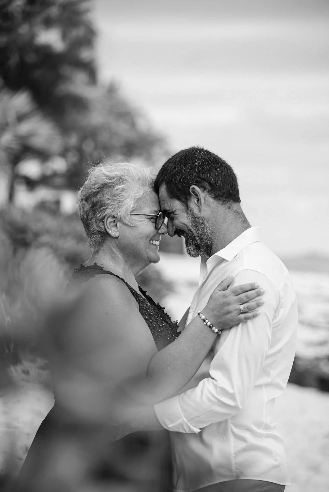 A joyful couple shares a loving moment outdoors, touching foreheads and smiling, with trees and a cloudy sky in the background.