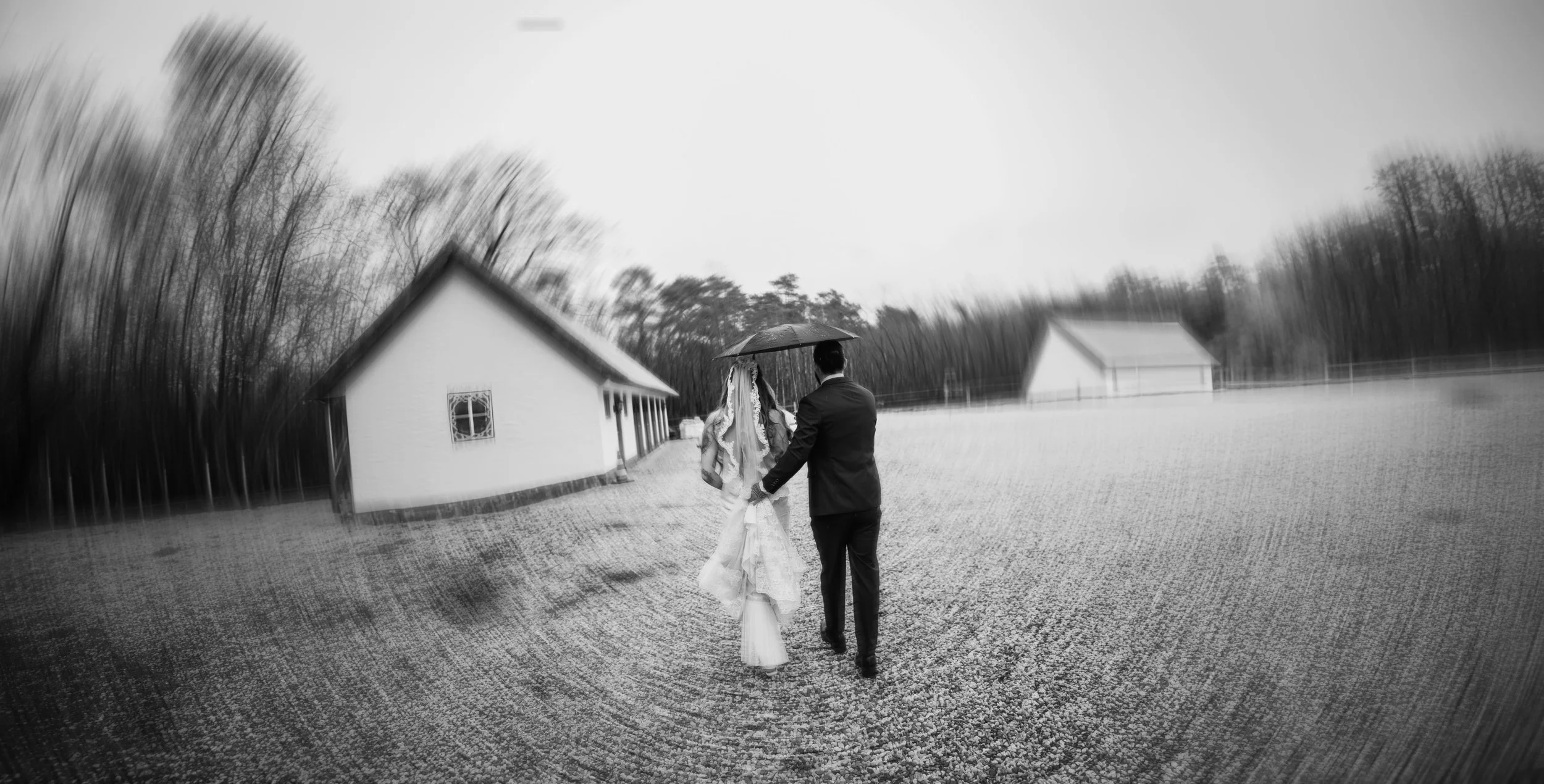 Black and white photo of a bride and groom walking together under an umbrella outside on a cloudy day, with houses and trees in the background.