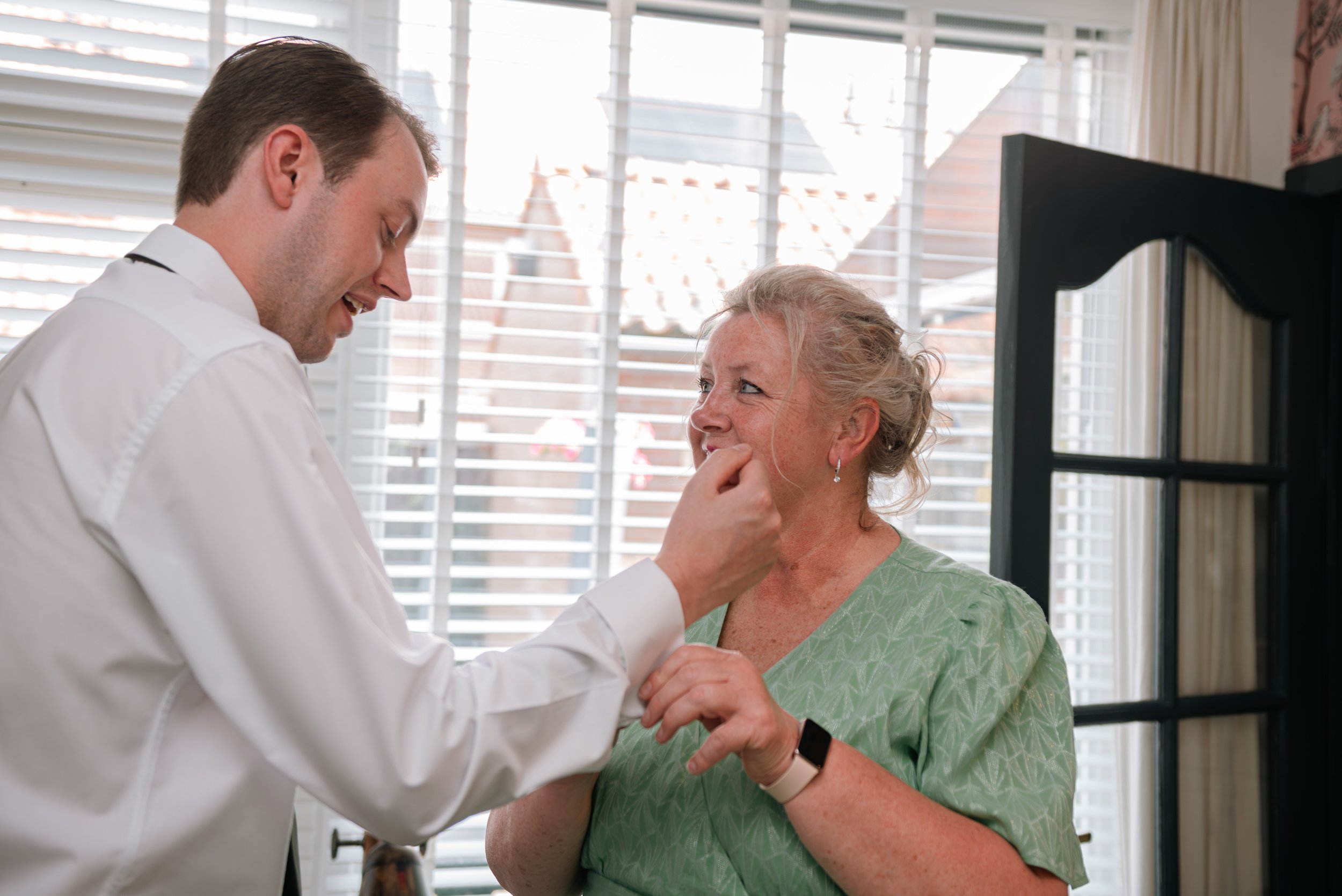 A man in a white shirt helping an elderly woman in a green hospital gown with a medical device on her wrist, indoors near a window with blinds.