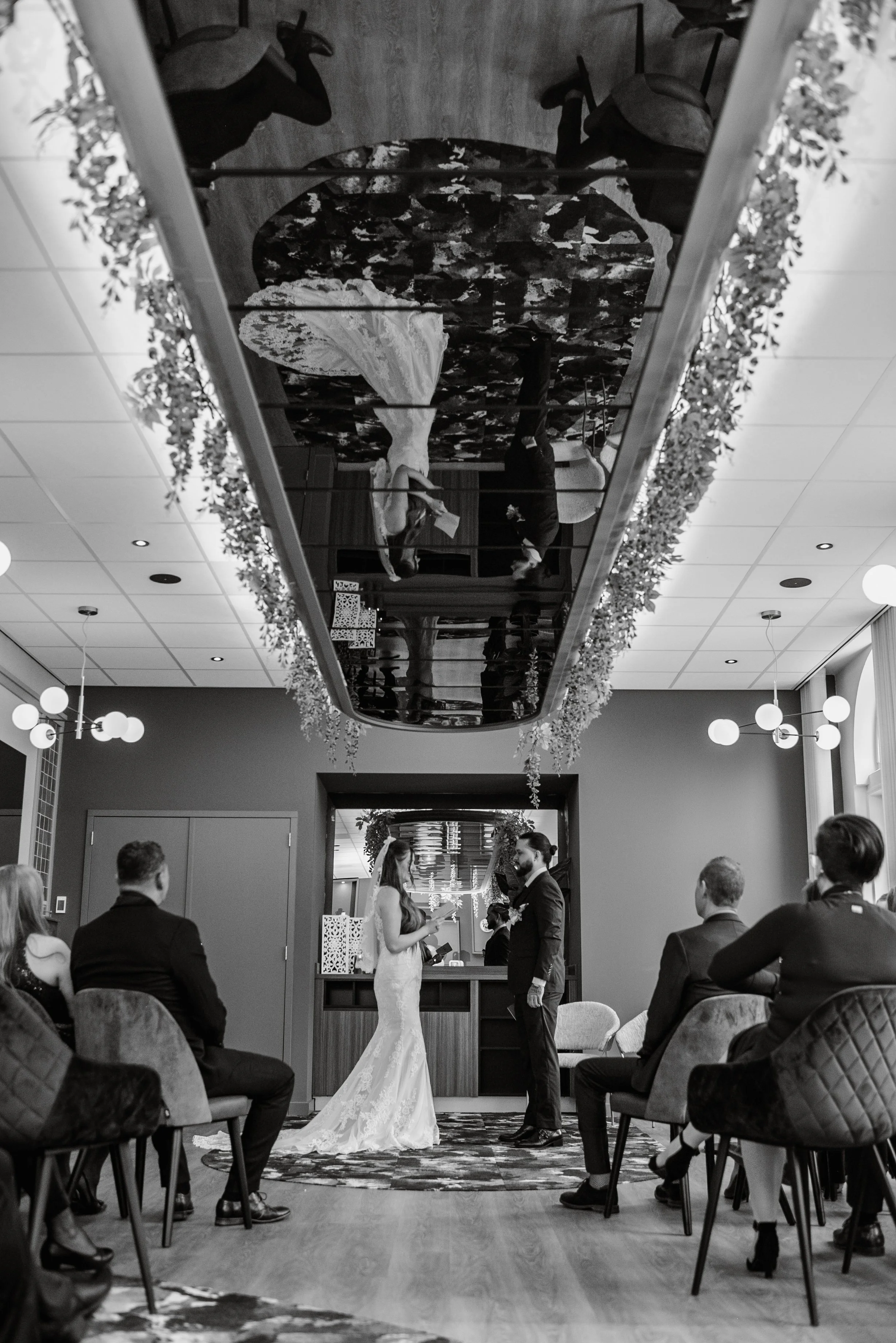 A wedding ceremony with a bride and groom standing in front of an officiant in a decorated indoor venue with guests seated on both sides.