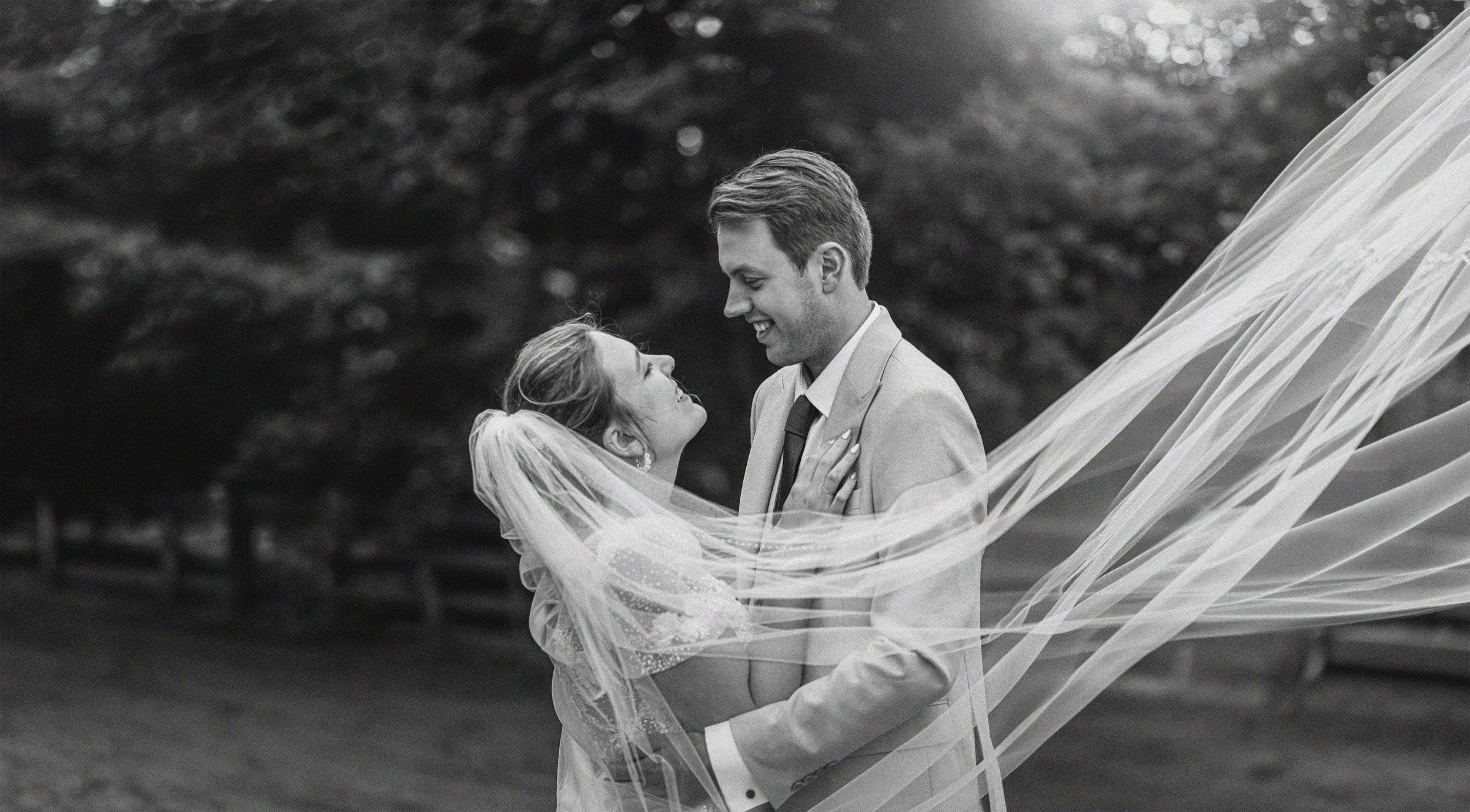 A black-and-white photograph of a bride and groom in a joyful moment. The groom, dressed in a suit, smiles while looking at the bride, who is wearing a wedding dress and veil. The bride is looking up at the groom with a happy expression, and her veil is flowing around them. There are trees in the background.