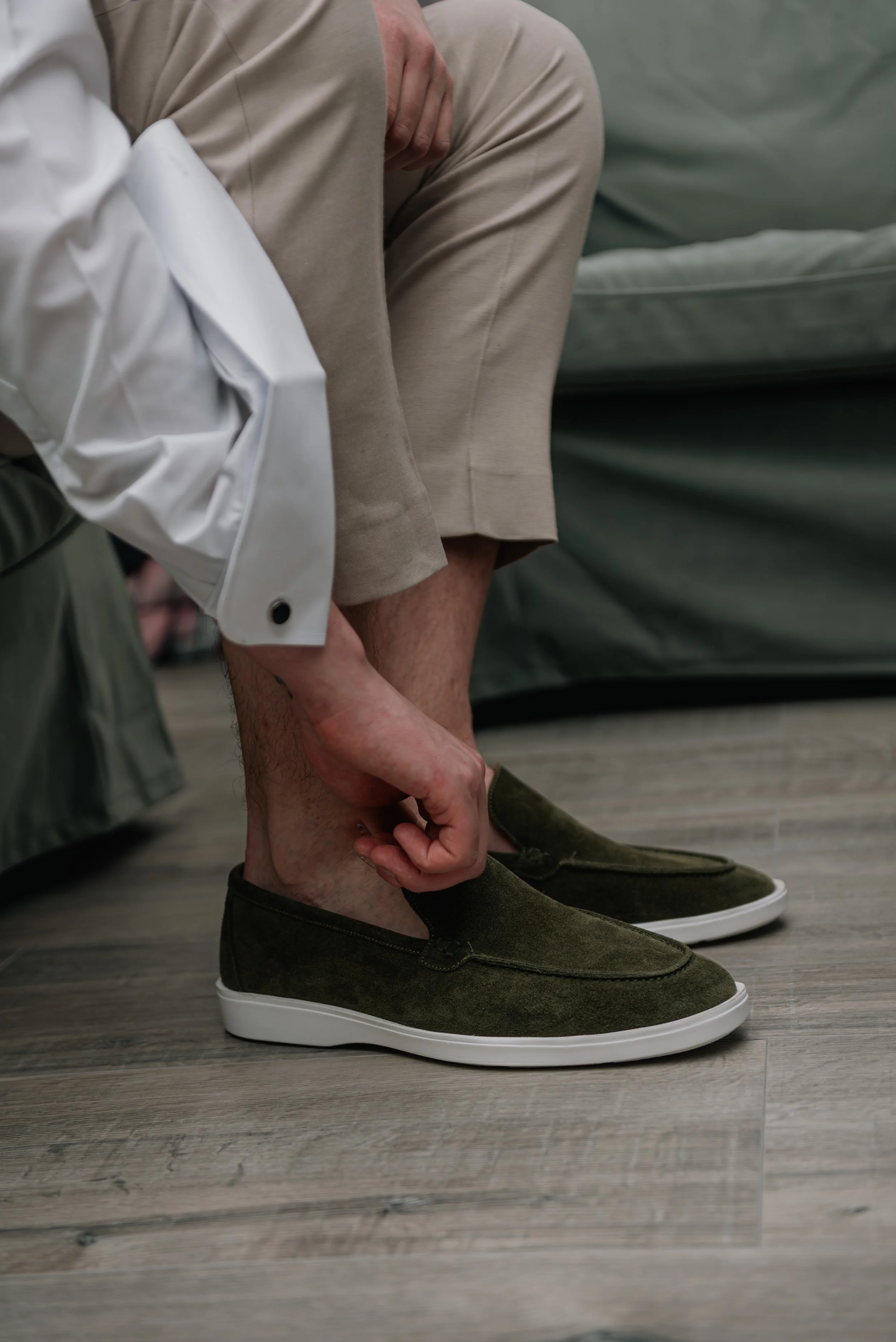 Person sitting with crossed legs, wearing beige pants and a white shirt, adjusting or tying their green sneakers on a wooden floor, with a gray sofa in the background.