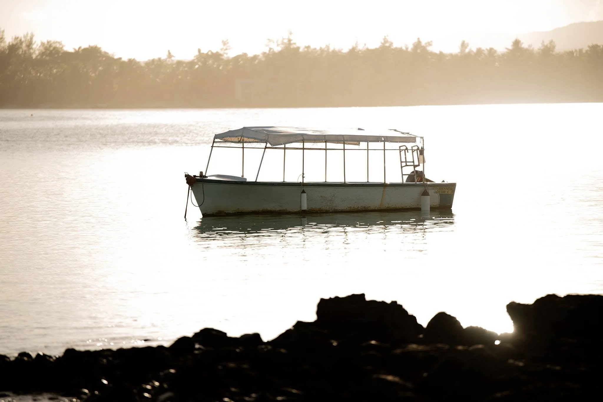 A boat floating on calm water during sunset, with a silhouette of rocks in the foreground and a tree-lined shoreline in the background.