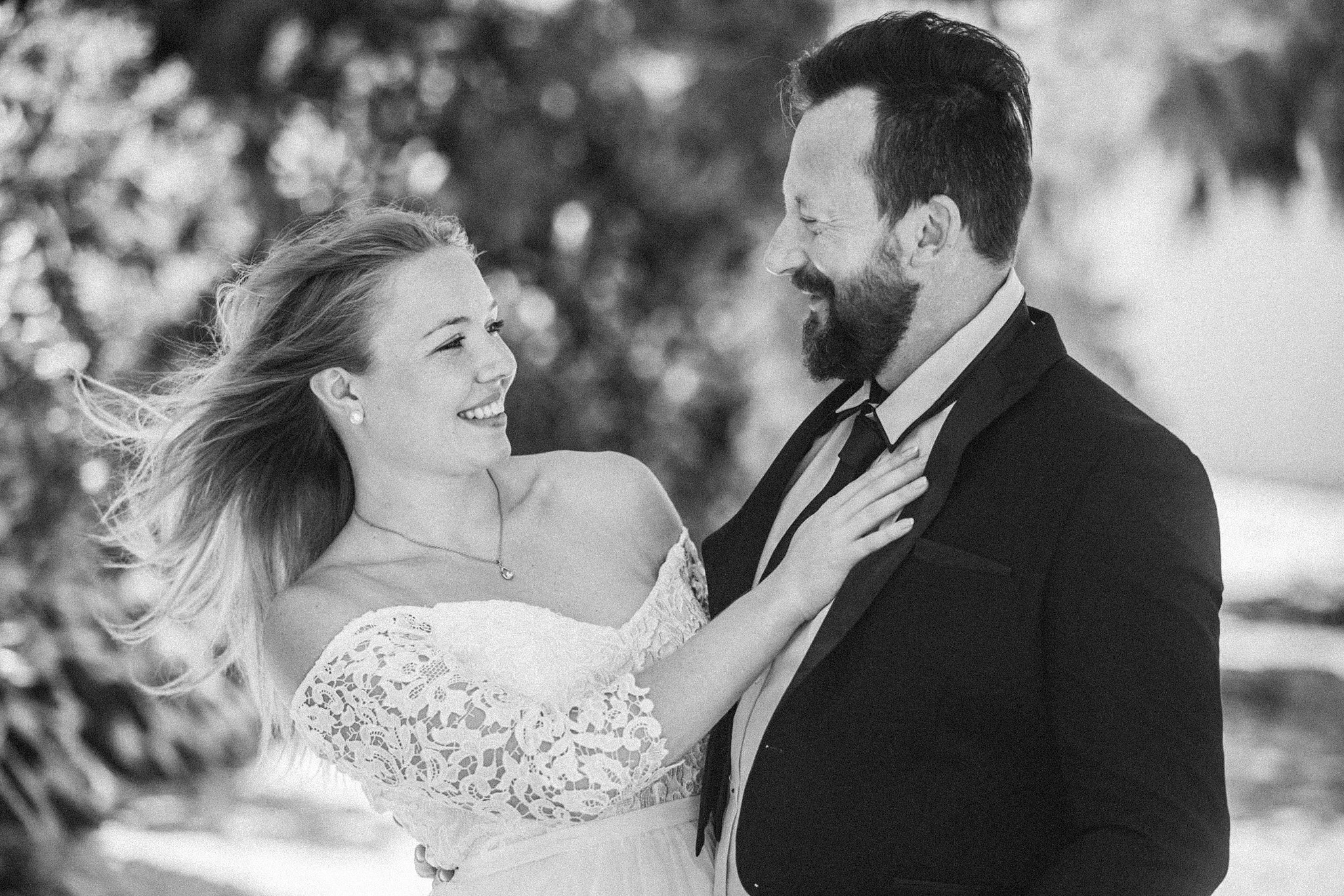 A smiling bride and groom dancing outdoors, looking at each other, with the bride wearing a lace wedding dress and the groom in a suit and tie.