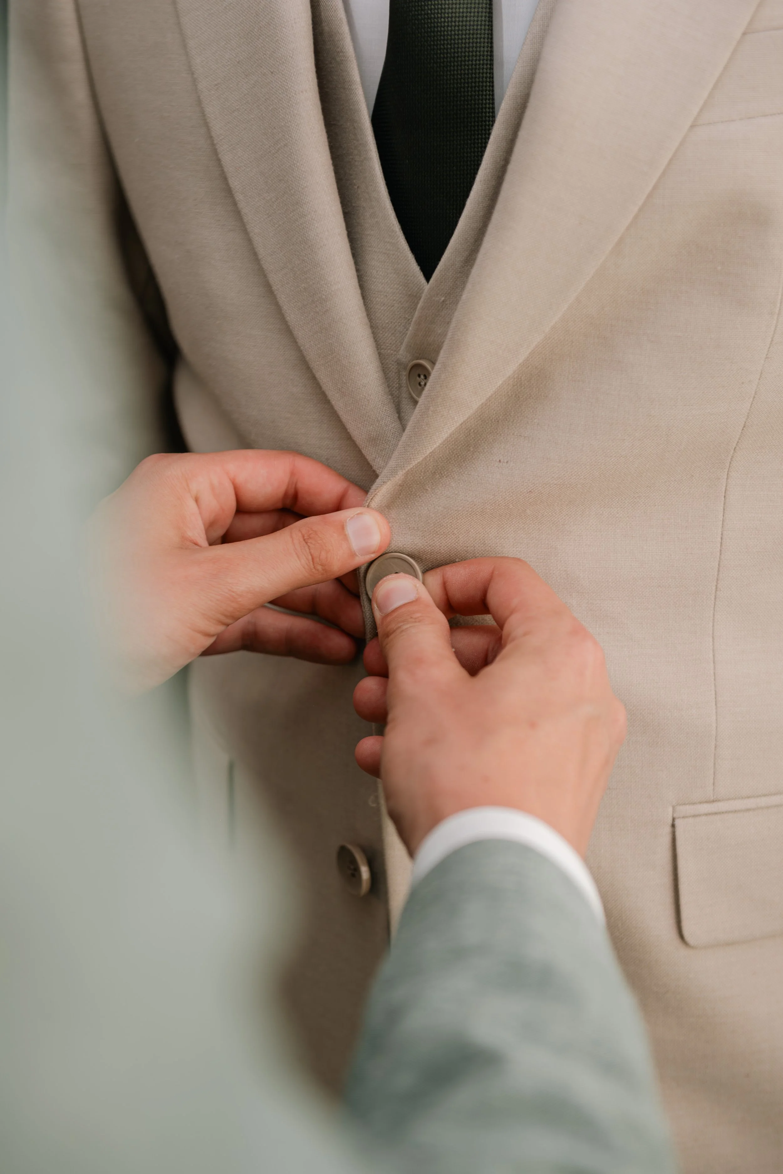 Close-up of a beige suit jacket being buttoned by a person in a grey suit with a white shirt, with a green background.