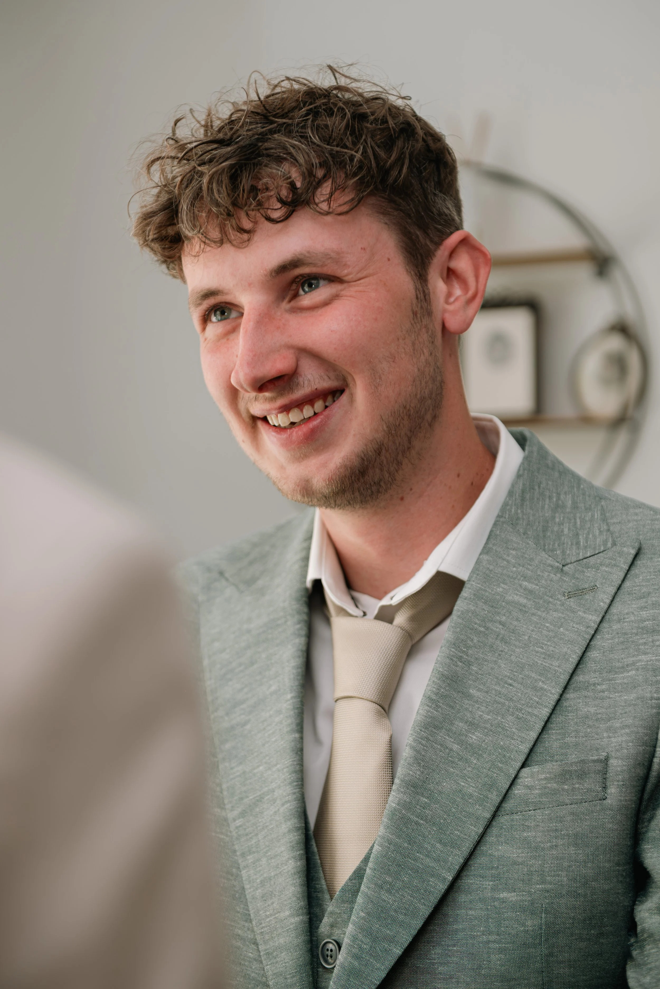 A smiling man with curly hair and a light beard wearing a gray suit and a beige tie, with a blurred background that includes a mirror and some fixtures.