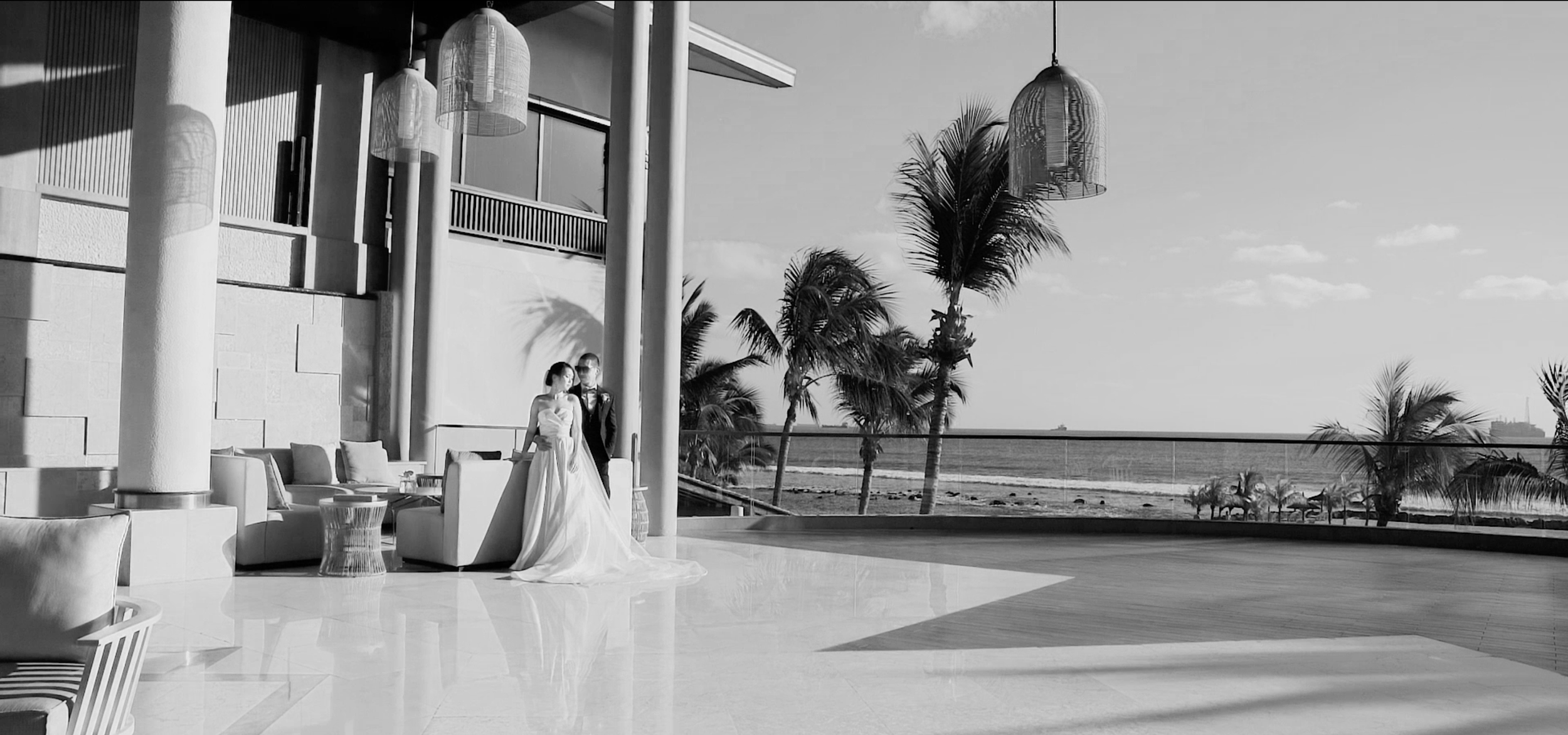 A black and white photo of a modern outdoor terrace at a beachfront resort. There are two people dressed in wedding attire, a bride and groom, standing near a sofa with decor cushions. The terrace offers a view of the sea, palm trees, and a distant ship or structure on the horizon. Large hanging lanterns and architectural columns are also visible.