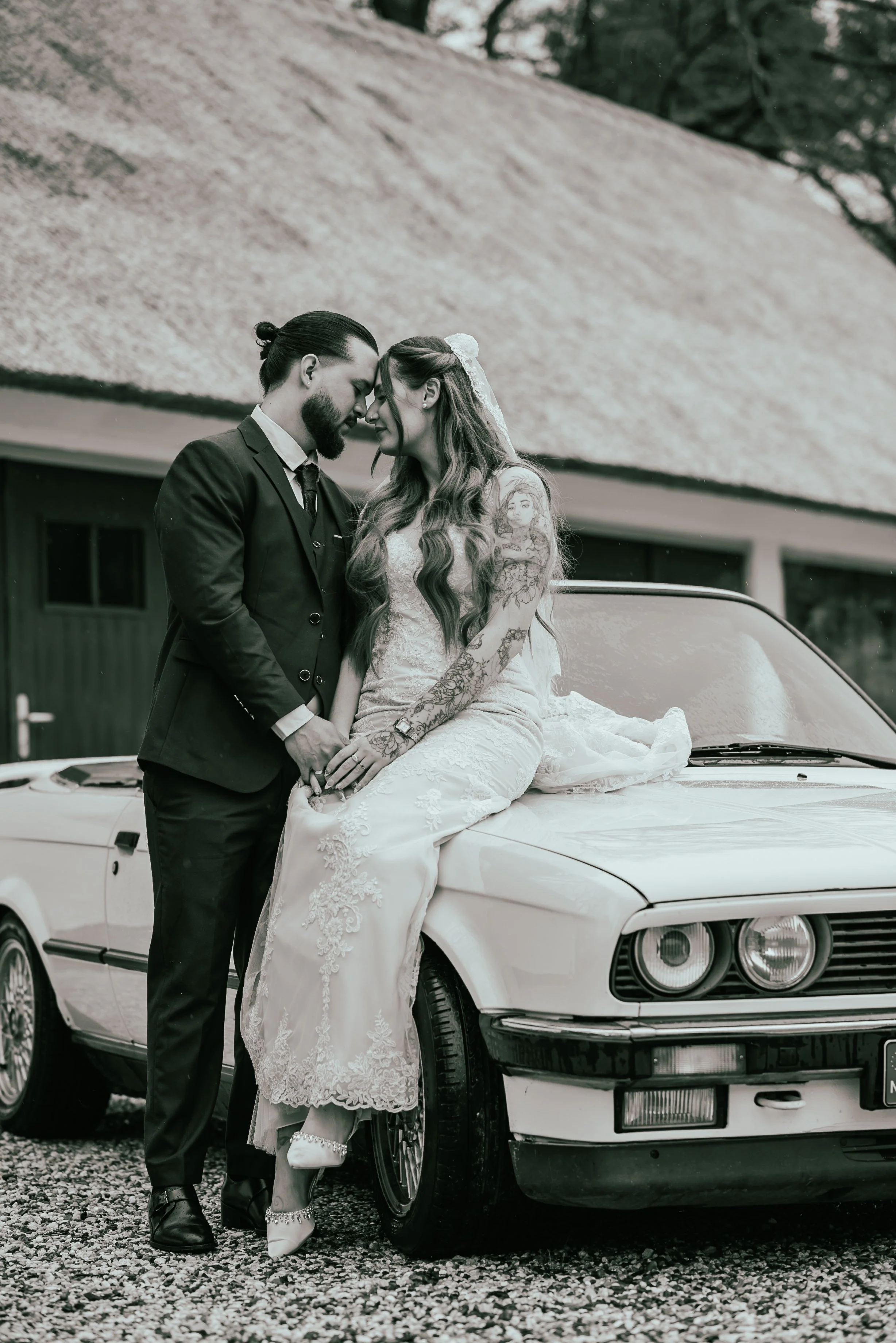 A couple dressed in wedding attire, sharing a forehead touch, leaning on a vintage car with a barn in the background.