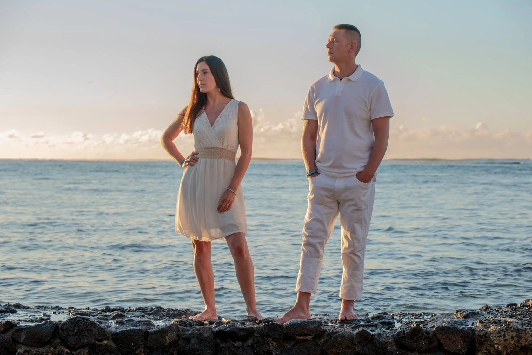 A man and woman standing on a rocky shoreline by the ocean at sunset, gazing into the distance with a calm expression.